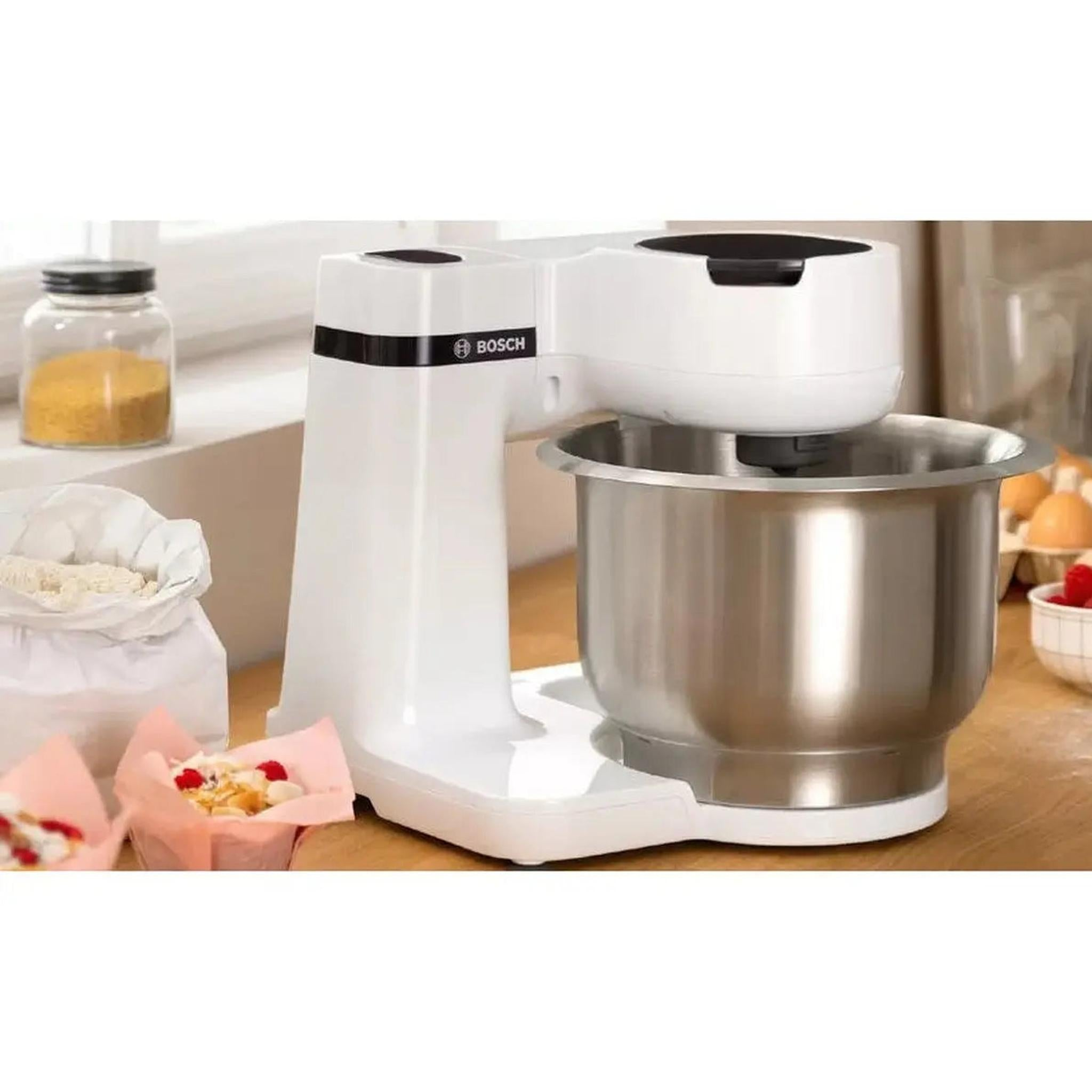 Een witte Bosch keukenmixer met een roestvrijstalen kom. Potten met ingrediënten, muffins en eieren op de achtergrond.
