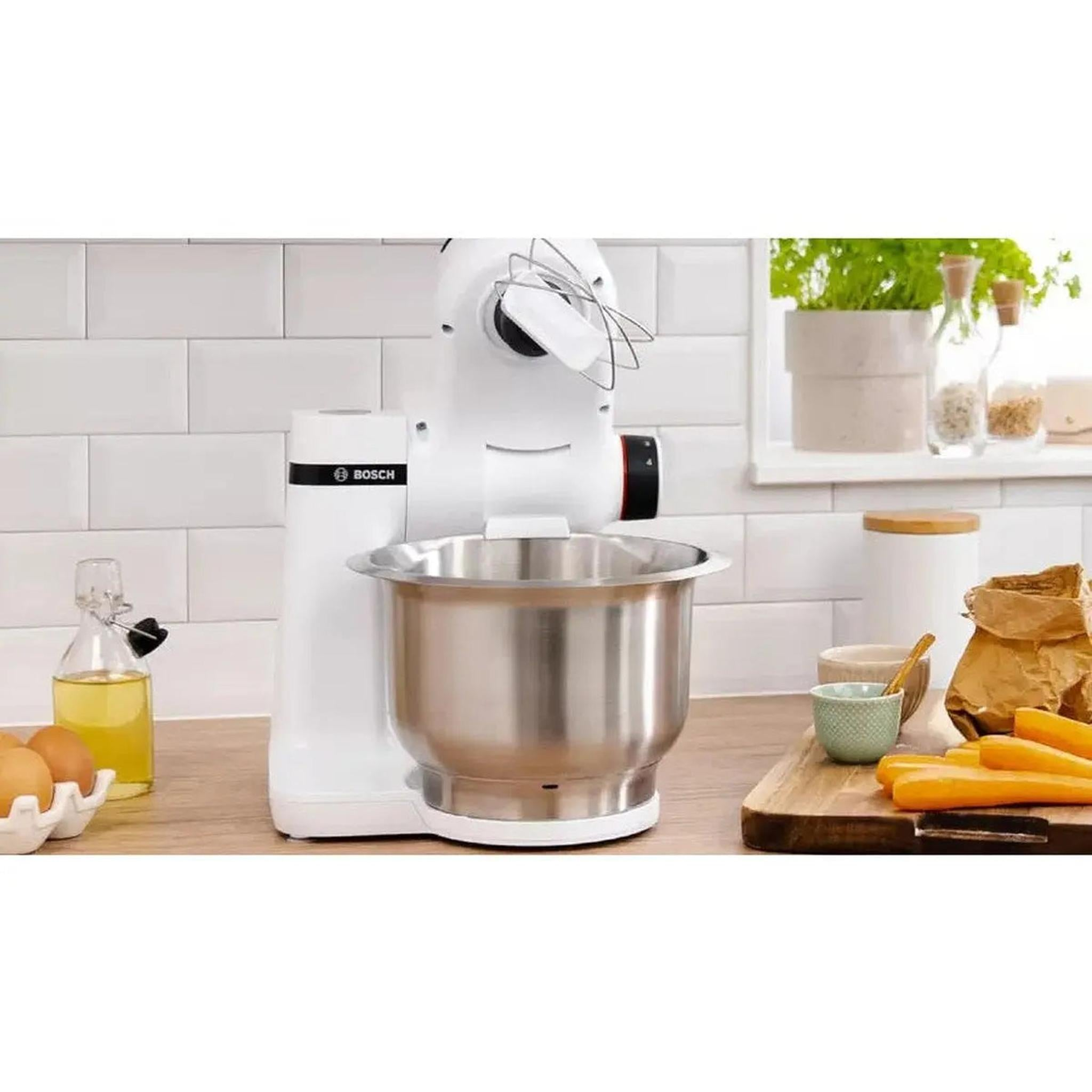 Een witte Bosch keukenmixer met een roestvrijstalen kom. Wortelen en eieren op het aanrecht. Een raam met planten op de achtergrond.