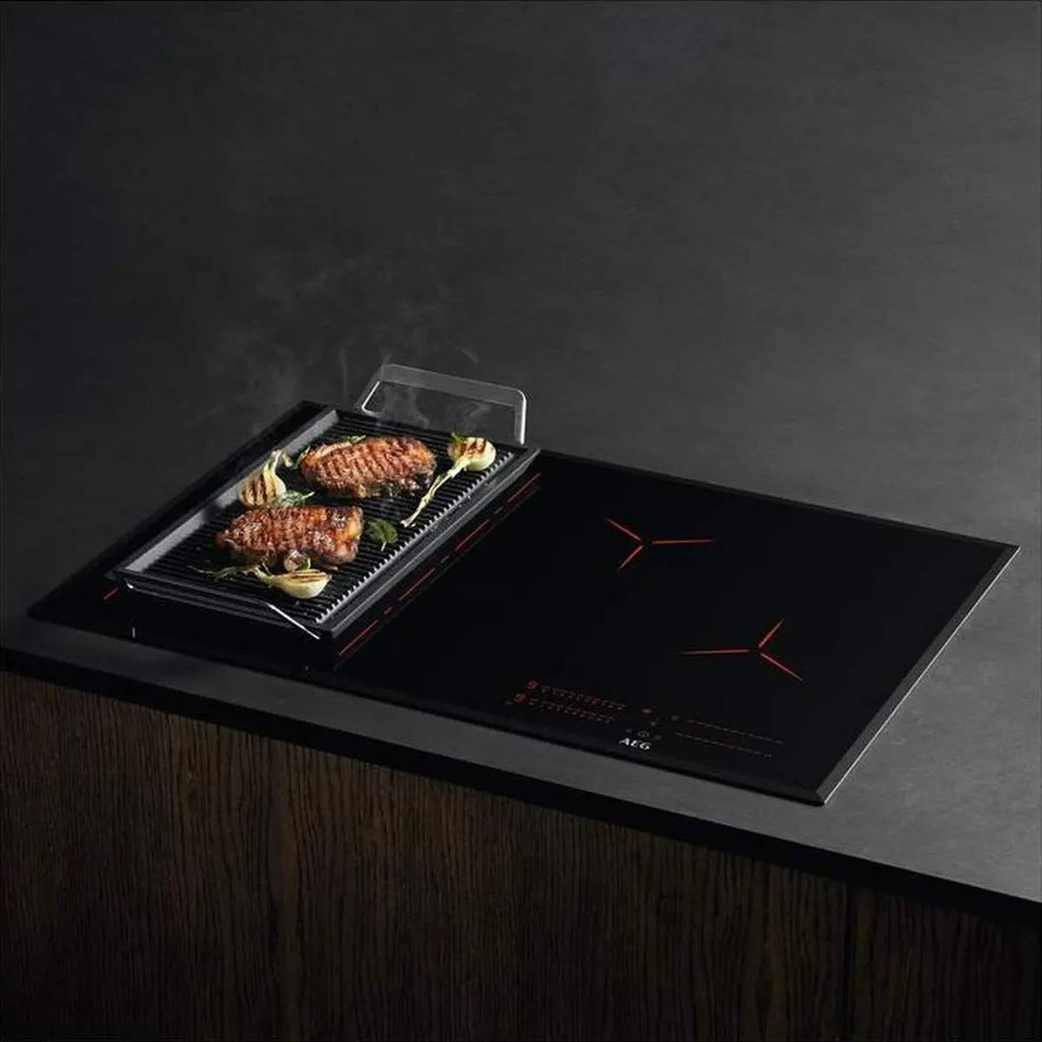 Zwart kookveld met grillpan. Twee steaks en groenten op de grill. AEG-logo. Houten kast eronder.