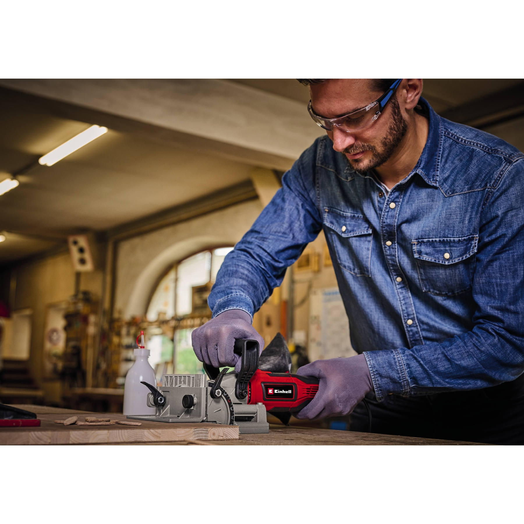 Ein Mann in einem Jeanshemd benutzt ein rotes Elektrowerkzeug auf Holz. Er trägt Handschuhe und eine Schutzbrille in einer Werkstatt.