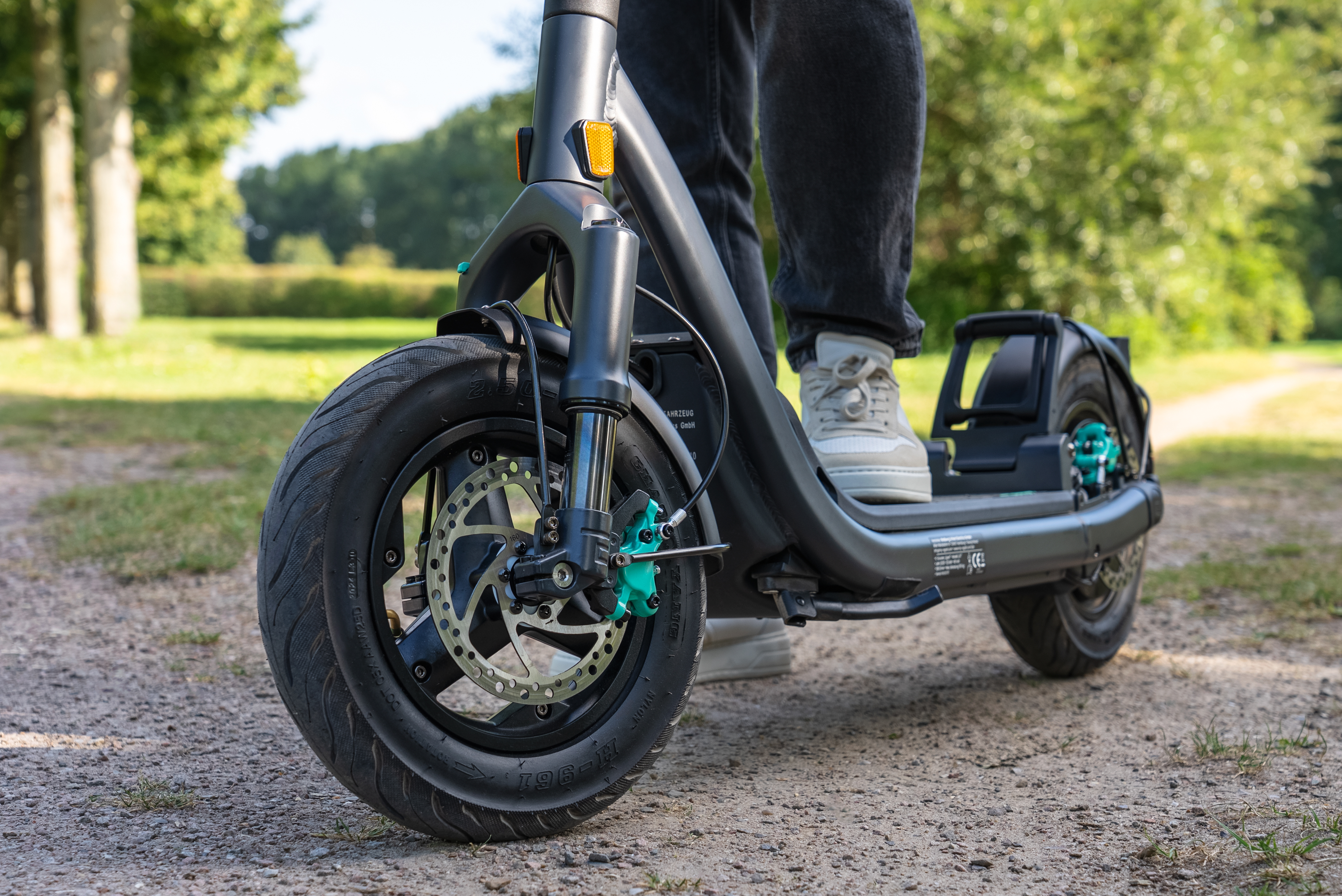 Eine Person auf einem grauen Elektroroller mit schwarzen Reifen auf einem Schotterweg. Grüne Bäume im Hintergrund.