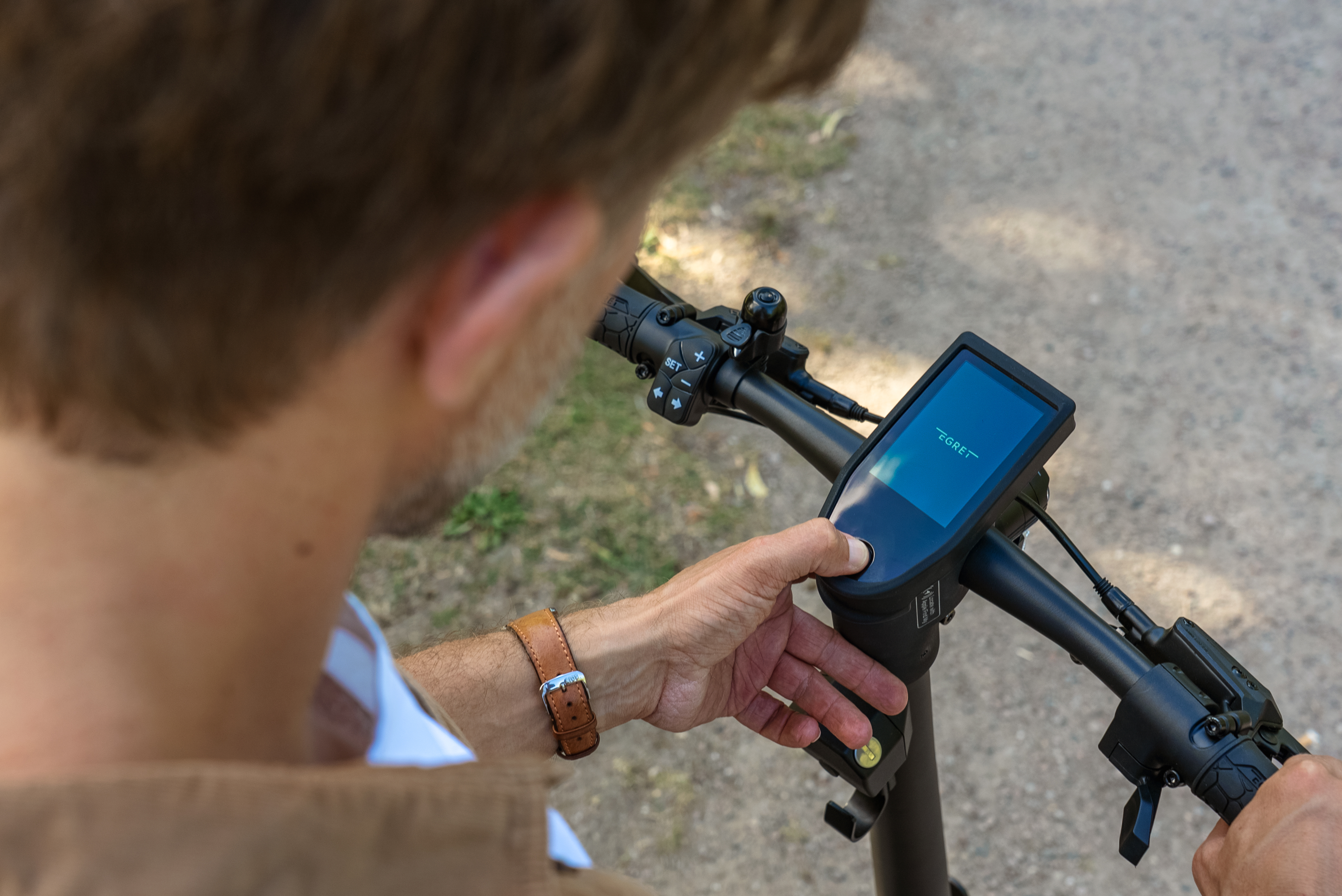 Person benutzt einen Elektroroller. Hand drückt die Power-Taste. Hintergrund ist ein verschwommener Pfad.