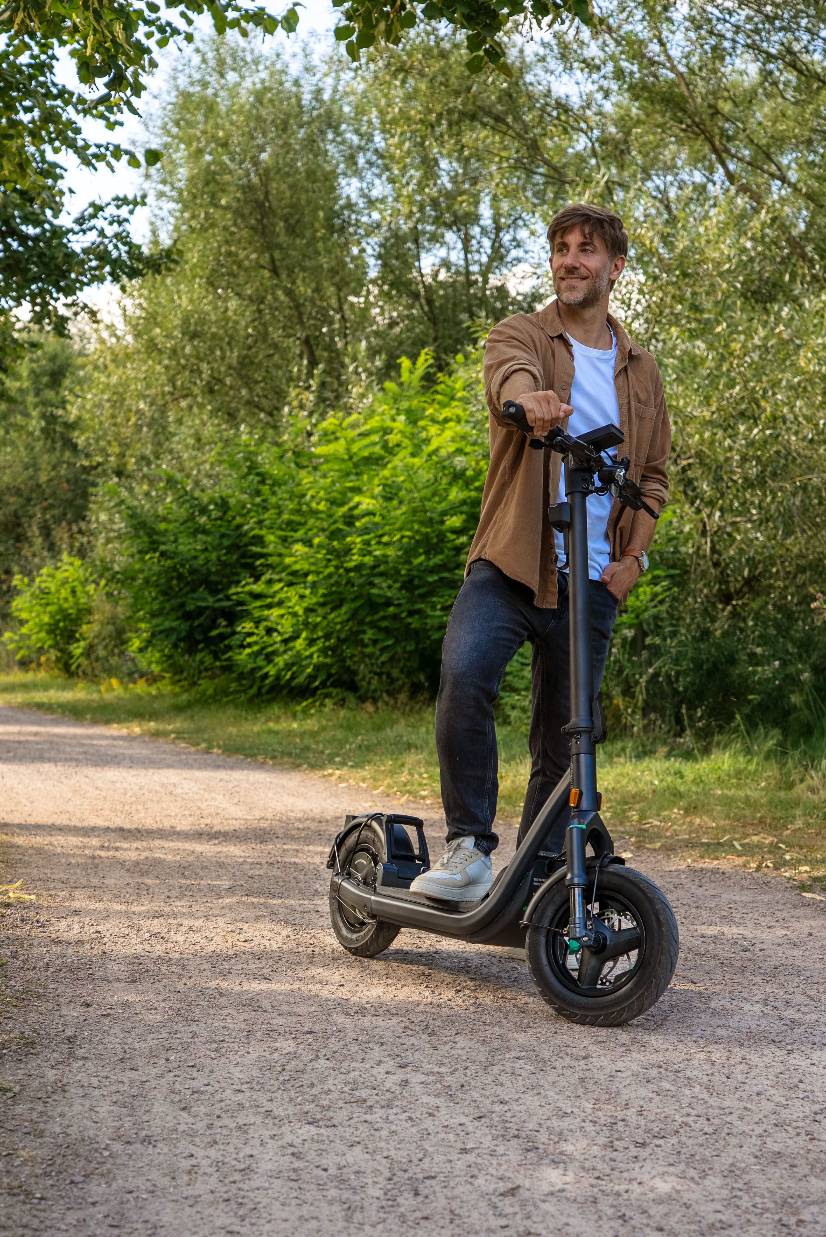Mann auf einem Elektroroller auf einem Schotterweg, umgeben von Bäumen. Er trägt ein braunes Hemd und Jeans.