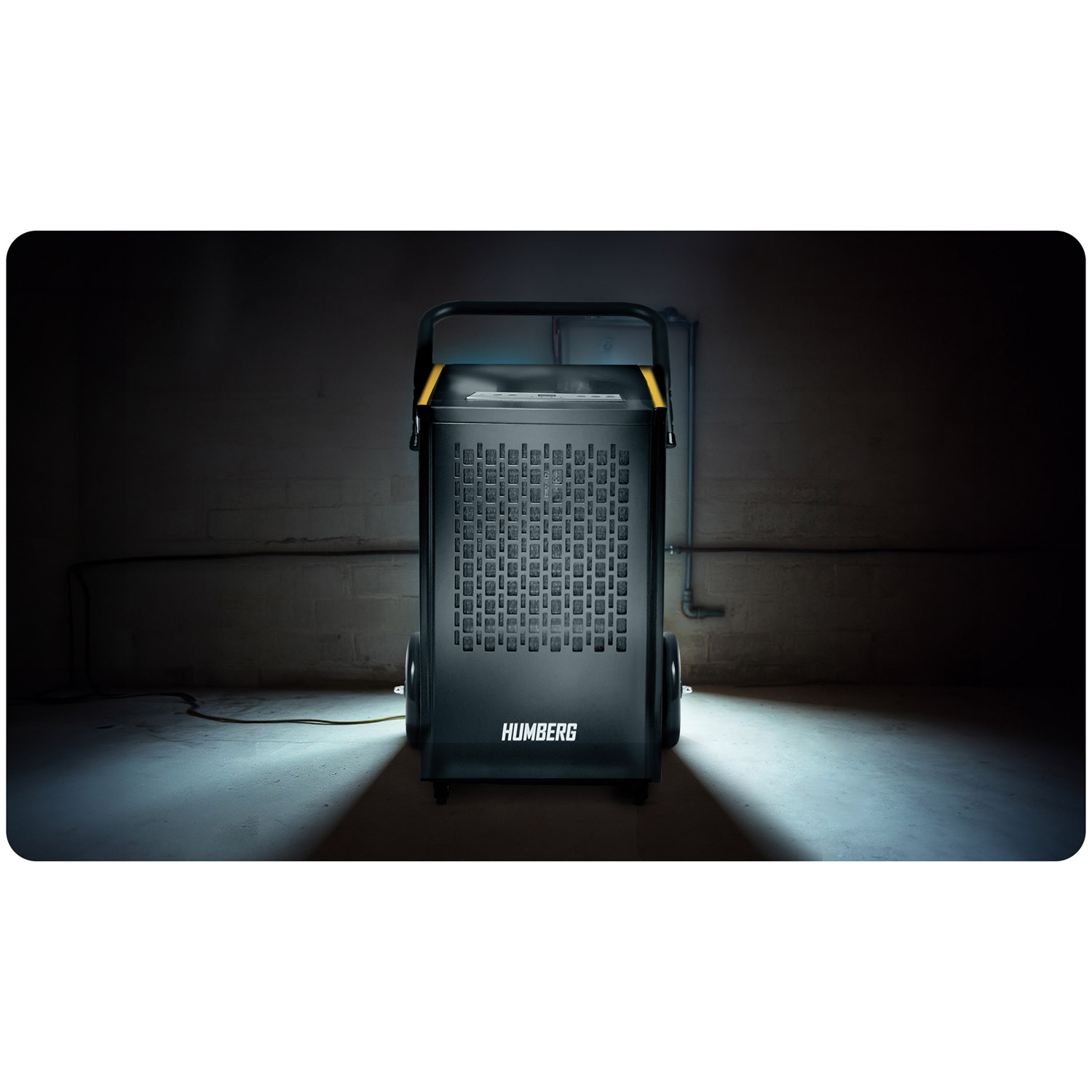 Odsuszacz Humberg stoi w słabo oświetlonej piwnicy. Jest czarny z wzorem kratki, kołami i uchwytem. Kabel biegnie w lewo.