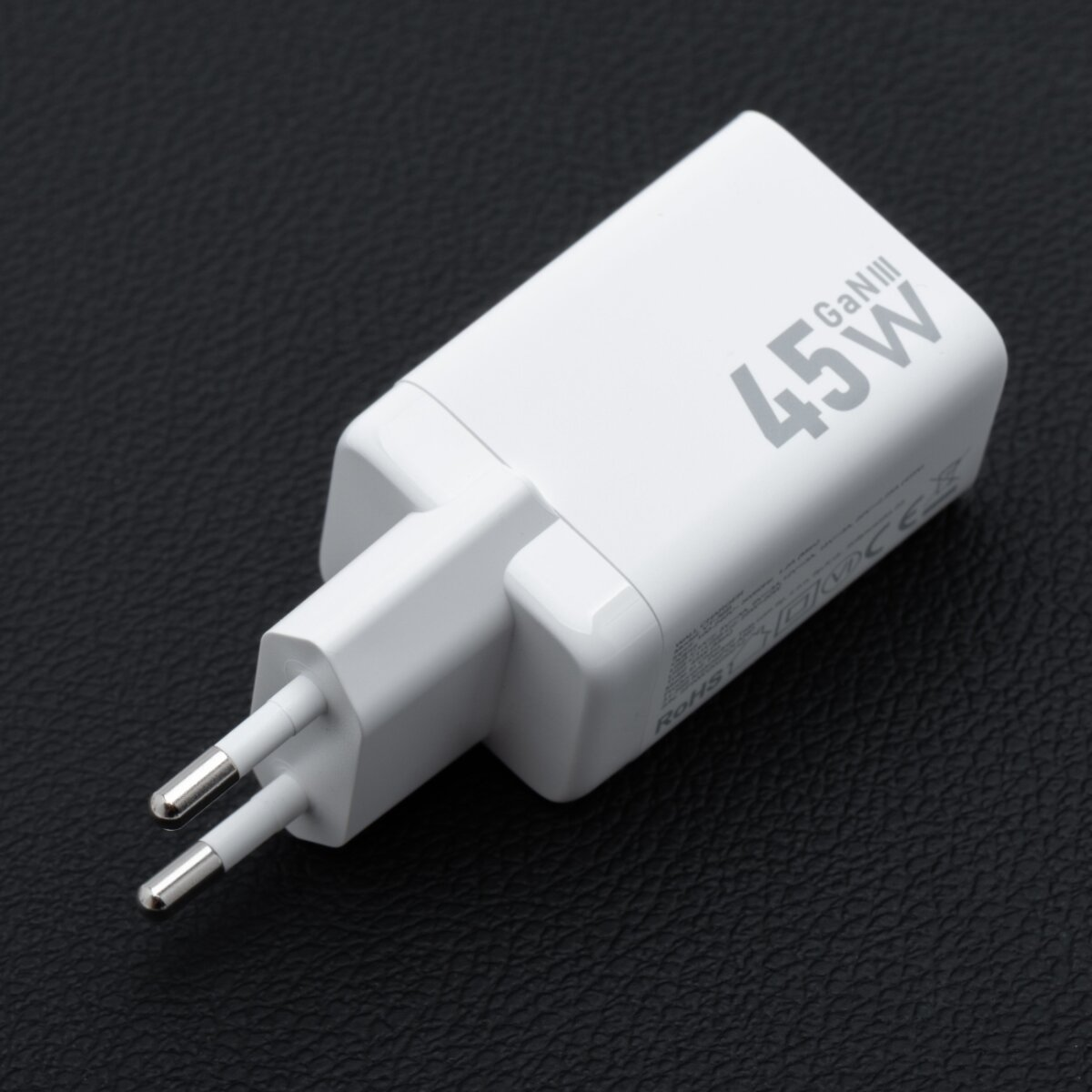 Un chargeur blanc GaNIII 45W sur une surface noire texturée. Le chargeur a deux broches métalliques et une étiquette imprimée.