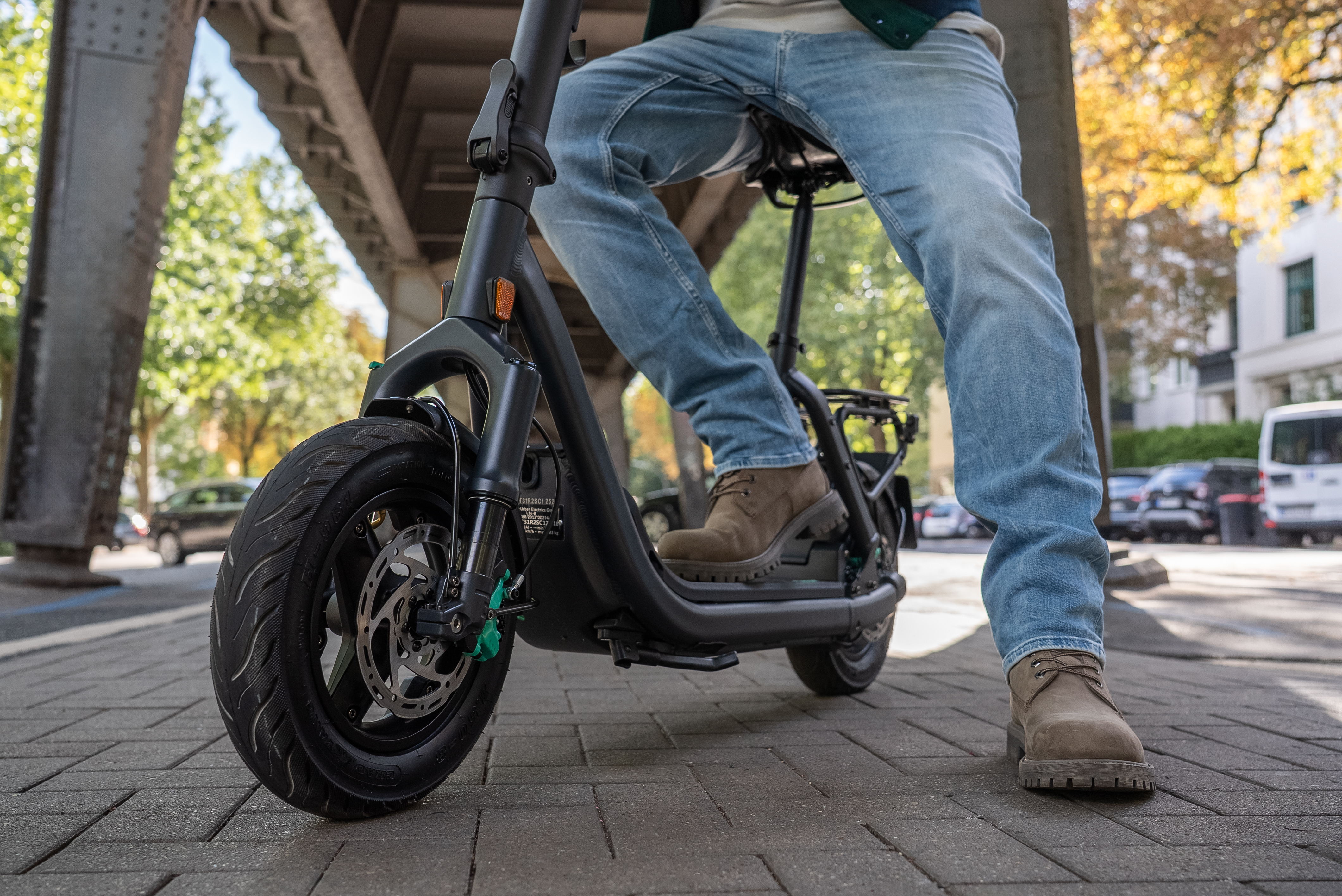 Person auf einem schwarzen Roller. Jeans und braune Stiefel. Bürgersteig und Gebäude im Hintergrund. Überführung oben.