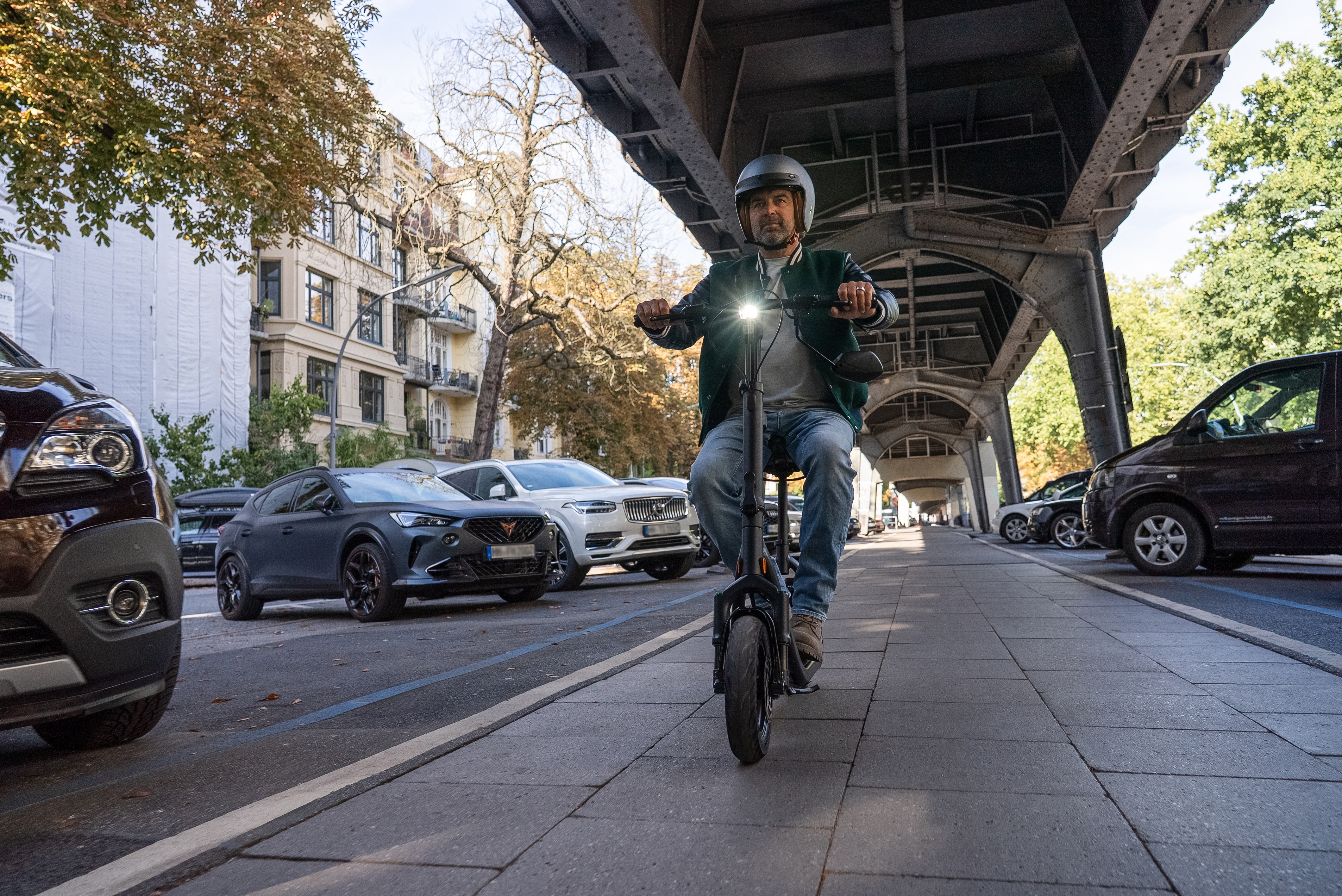 Mann auf E-Scooter unter einer Brücke. Autos und Gebäude im Hintergrund. Helm und grüne Jacke sind sichtbar.