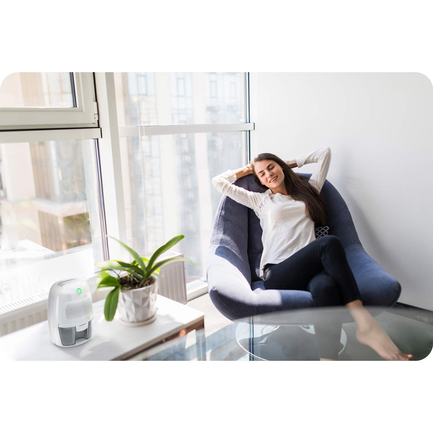 Kobieta relaksuje się w niebieskim fotelu, ręce za głową. Osuszacz powietrza i roślina stoją na stole przy oknie.