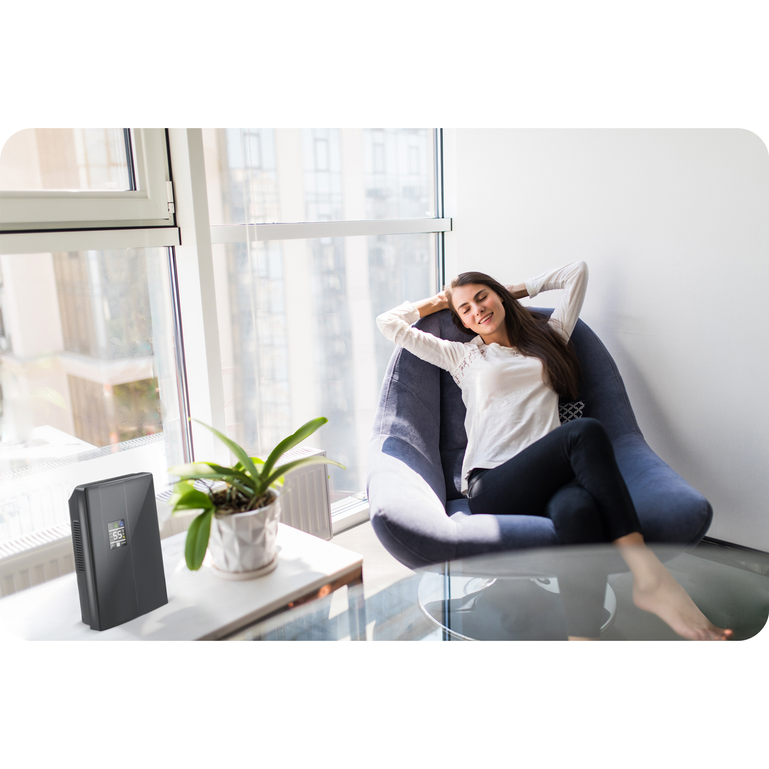 Kobieta relaksuje się w fotelu przy oknie. Osuszacz powietrza i roślina stoją na stole. Ma ręce za głową.