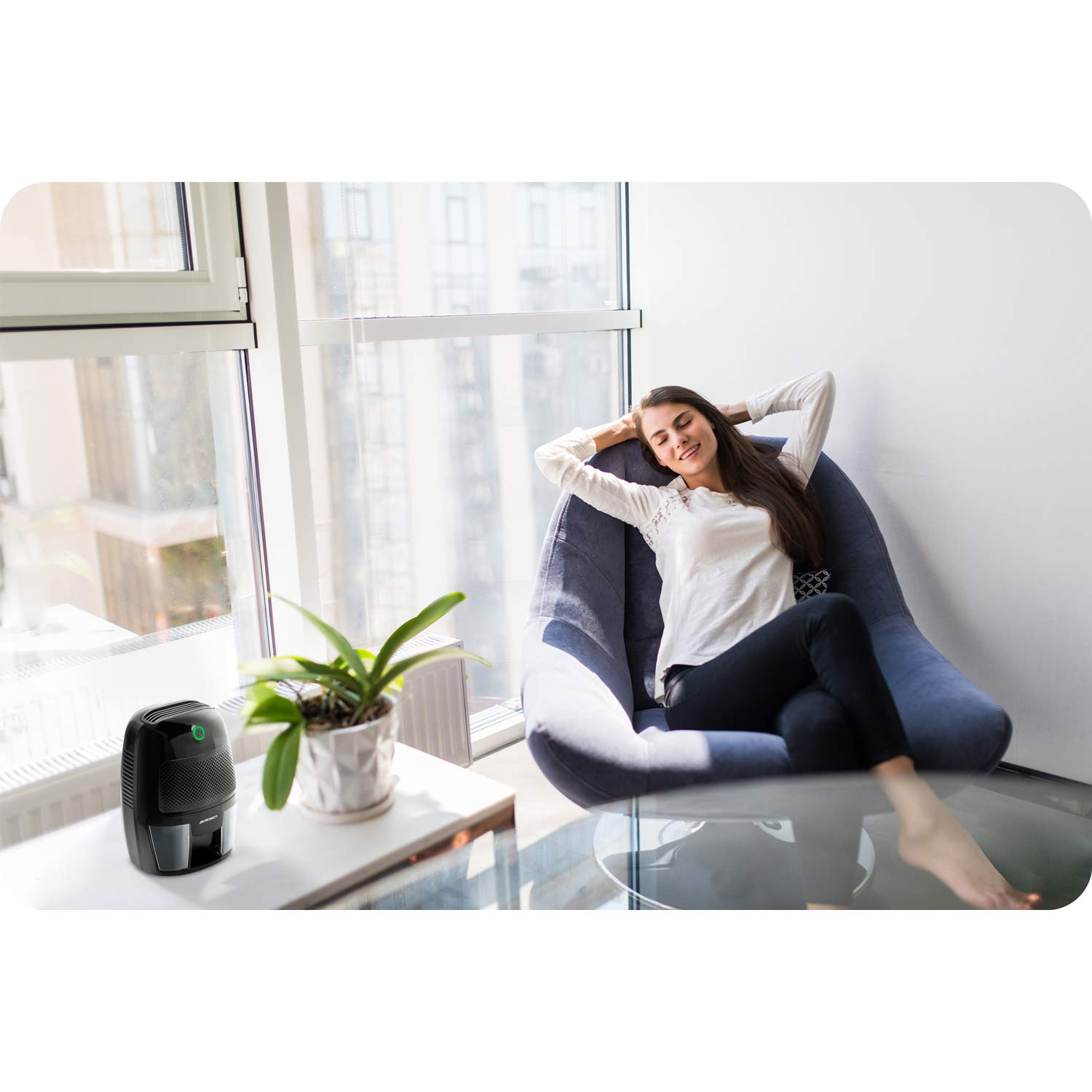 Kobieta relaksuje się na krześle, ręce za głową. Osuszacz powietrza i roślina na stole blisko okna. Słoneczny dzień.
