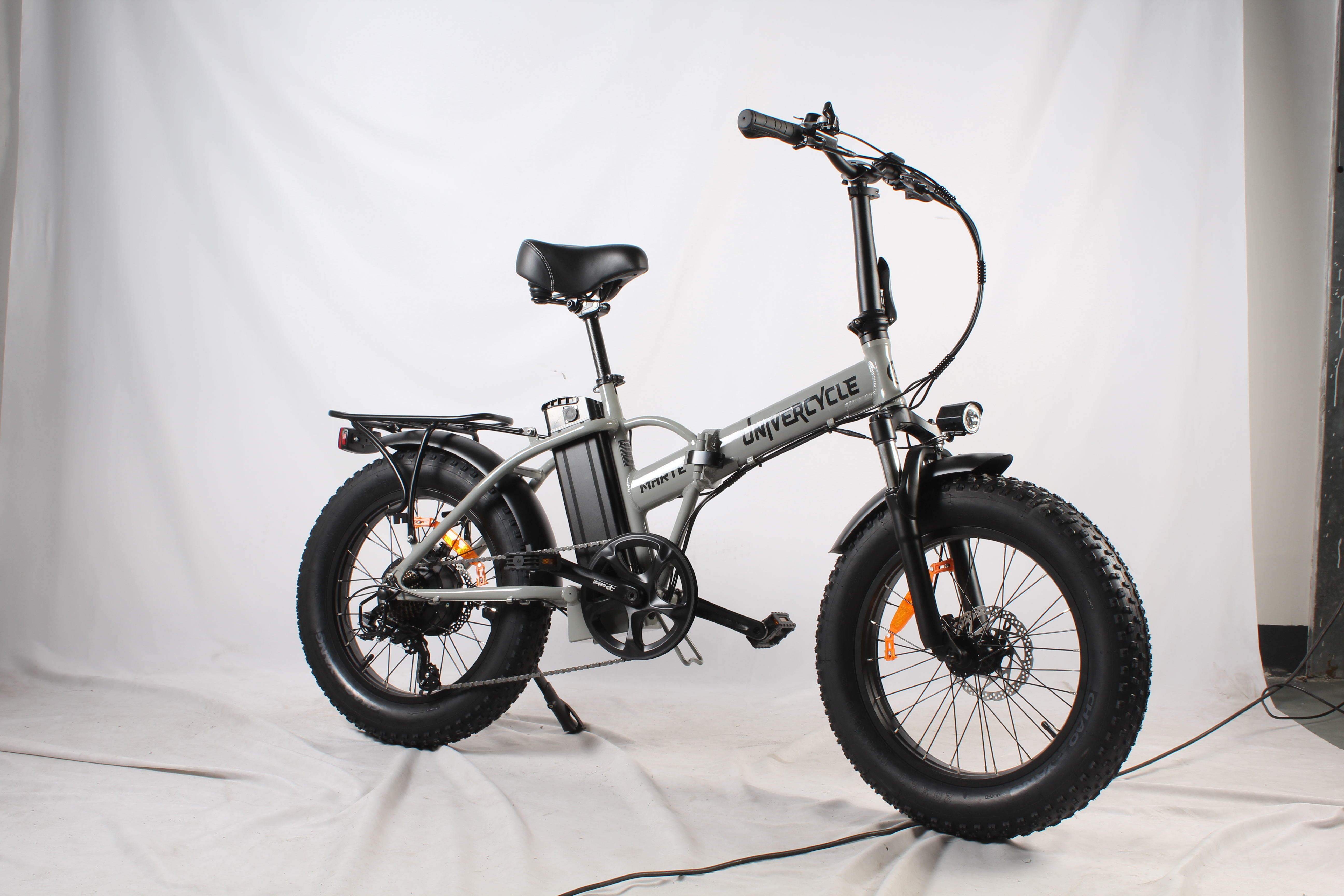 Una bicicletta elettrica grigia e nera. Ha pneumatici larghi, un sedile nero e un portapacchi. Lo sfondo è bianco.