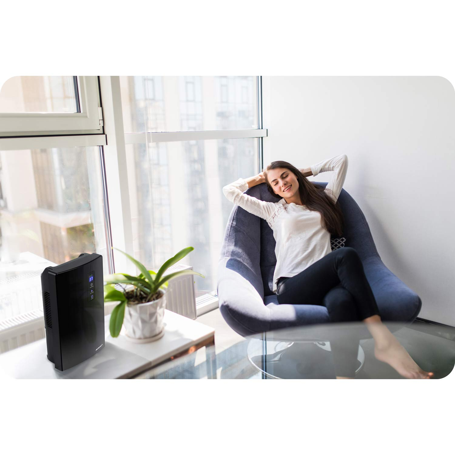 Kobieta odpoczywa na krześle. Widoczny jest czarny oczyszczacz powietrza, roślina i okno. Pokój jest jasny i przestronny.