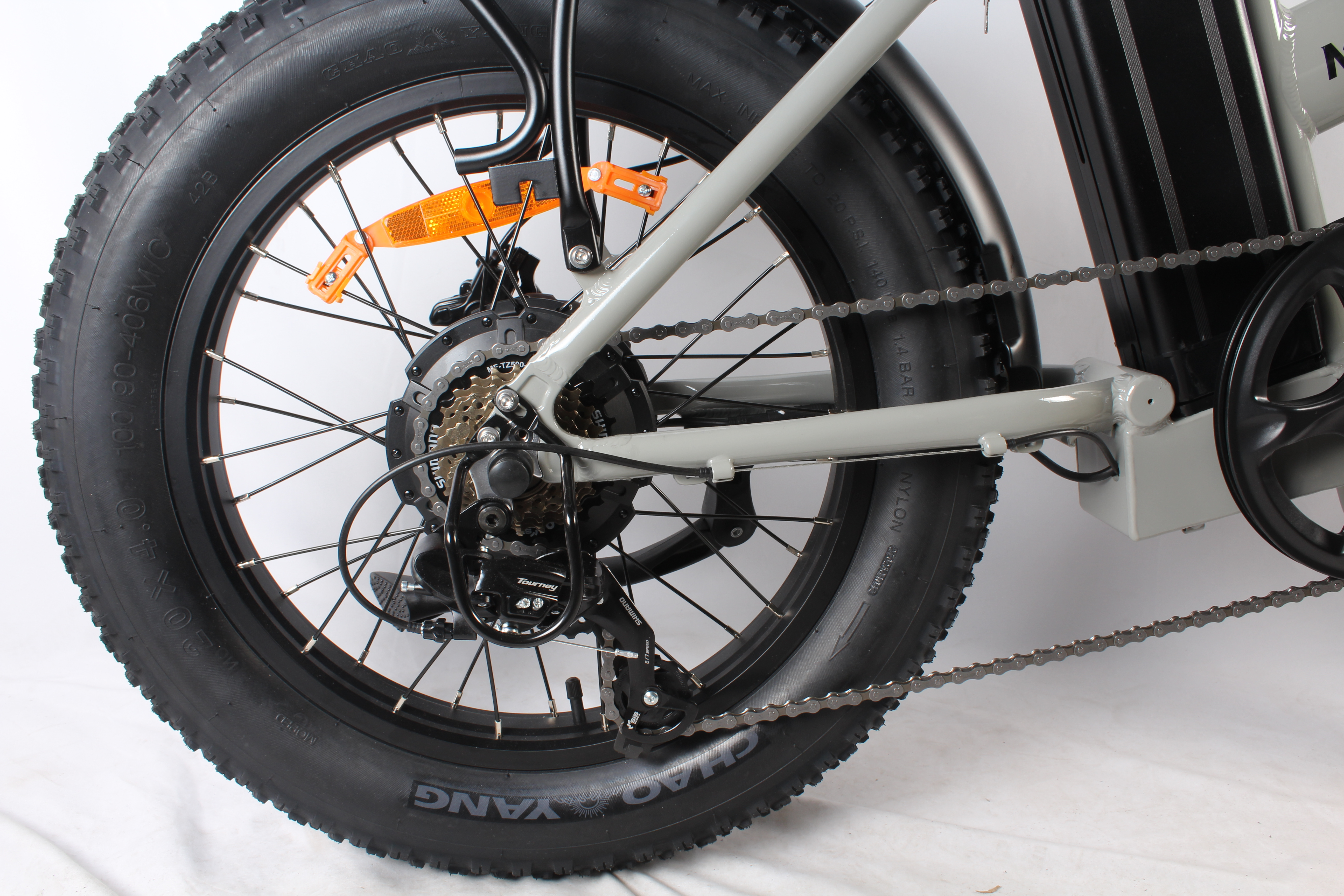 Primo piano di uno pneumatico nero di bicicletta con telaio grigio. Catena e ingranaggi visibili. Catadiottri arancioni sui raggi.