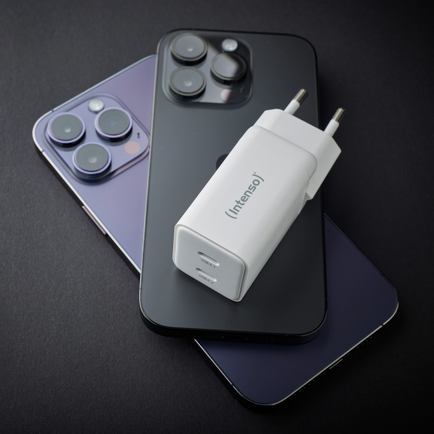 Drei Telefone, ein weißes Ladegerät auf schwarzer Oberfläche. Telefone sind schwarz, lila und dunkelblau. Ladegerät hat USB-C-Anschlüsse.
