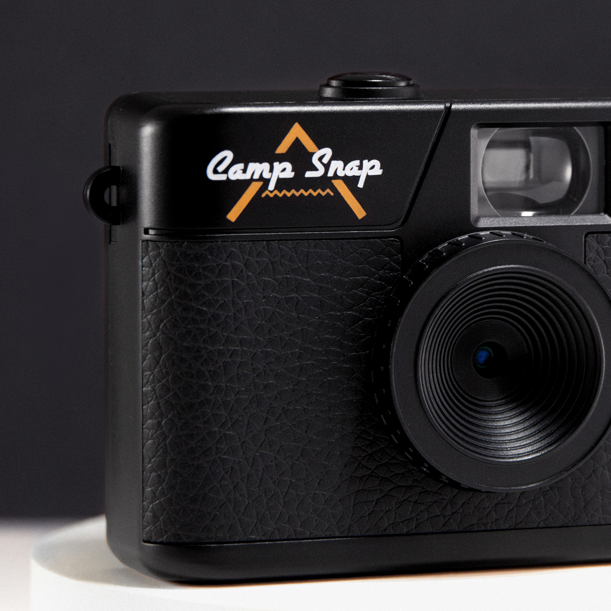 Schwarze 'Camp Snap'-Kamera. Weißer Text oben, orangefarbenes Berglogo. Strukturierte Vorderseite, Linse in der Mitte, schwarzer Hintergrund.