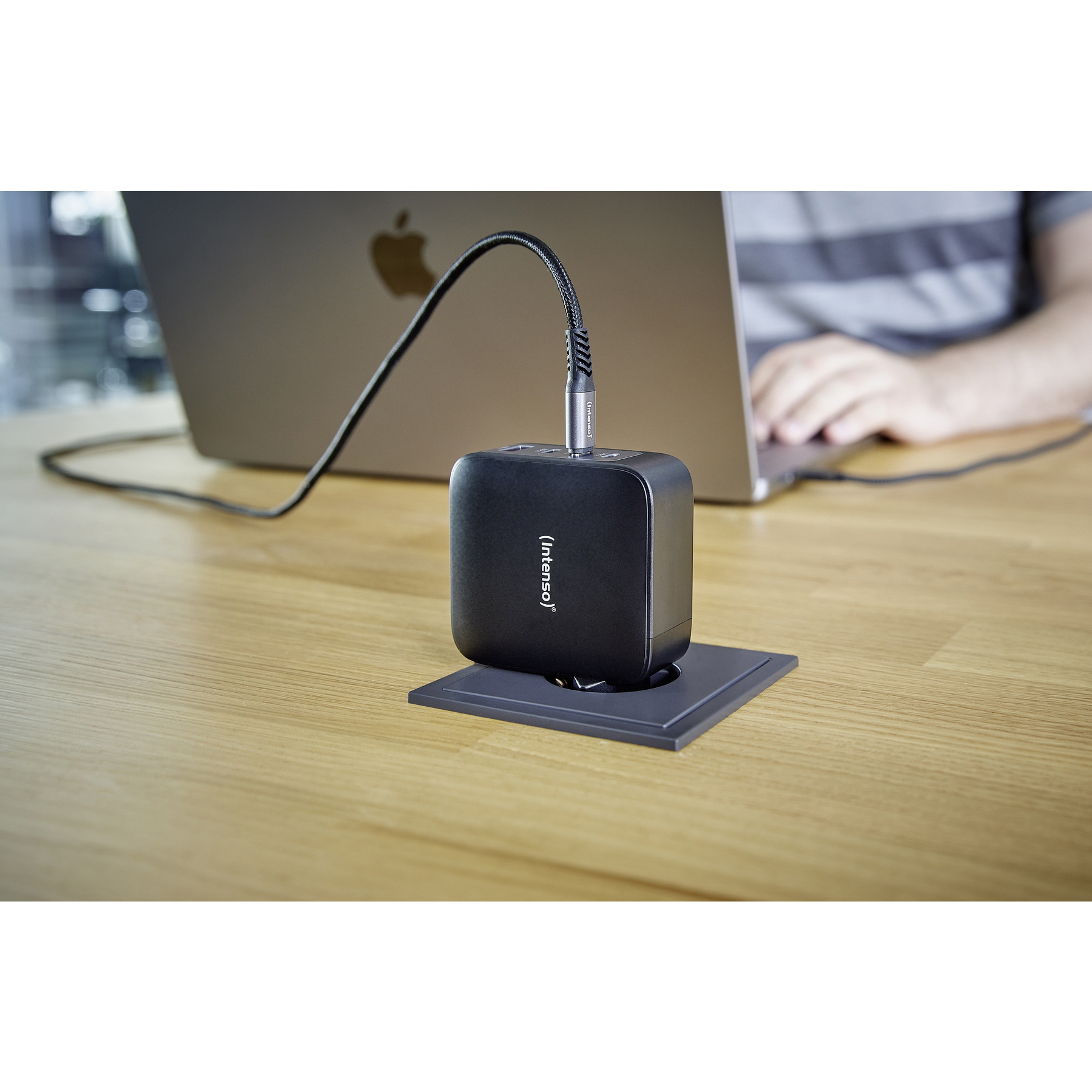 A black Intenso charger is plugged into a MacBook. The charger sits in a recessed outlet on a wooden desk. A person types.