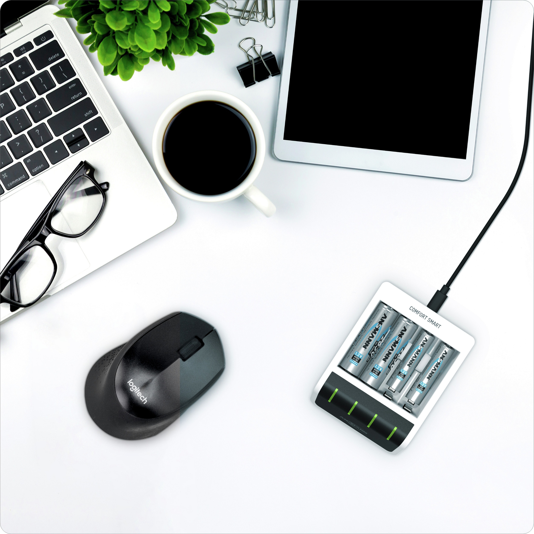 Spazio di lavoro con laptop, caffè, occhiali, mouse, caricatore con batterie, tablet e pianta su superficie bianca.