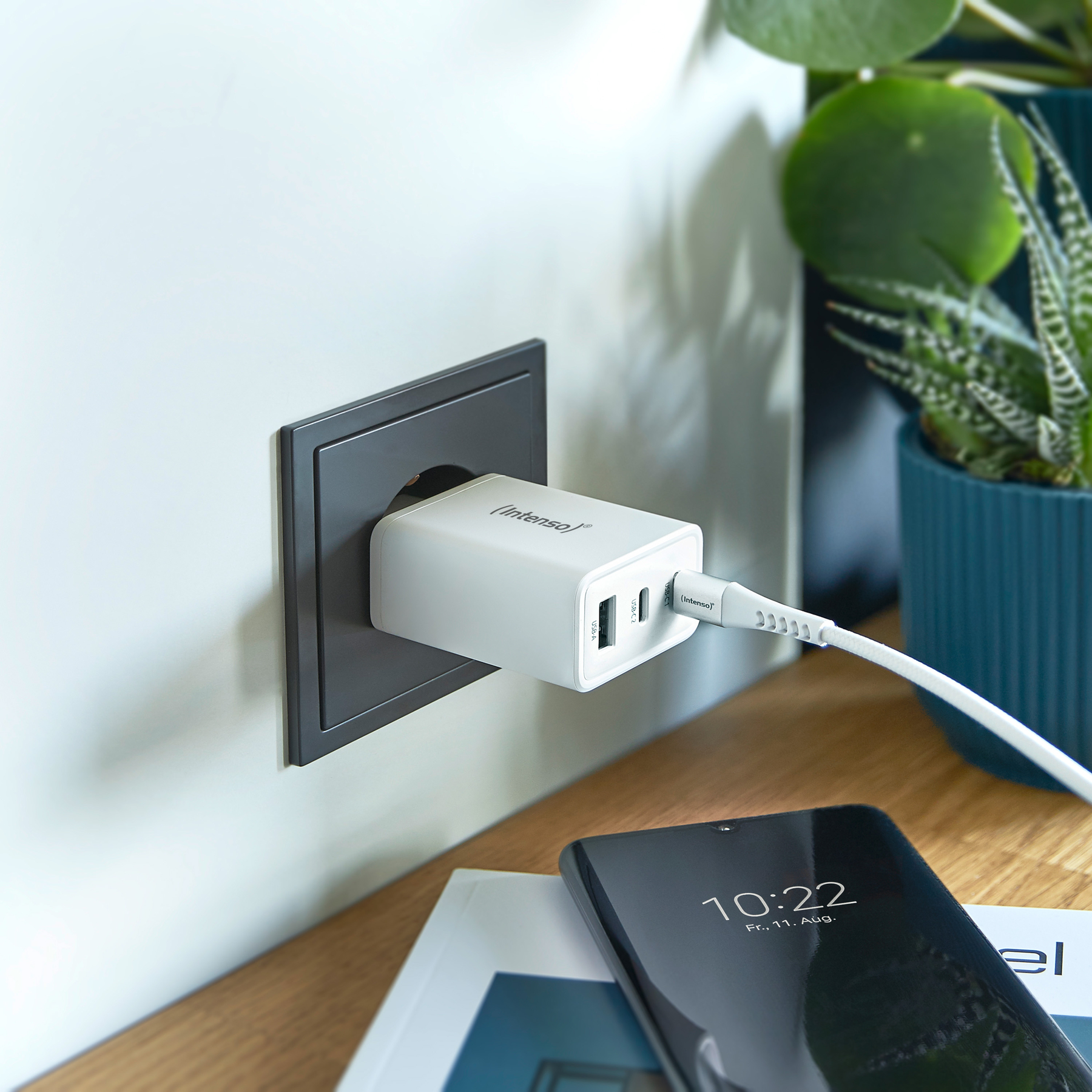Un cargador blanco enchufado a un enchufe. Un teléfono que muestra las 10:22 está sobre un escritorio. Plantas en el fondo.