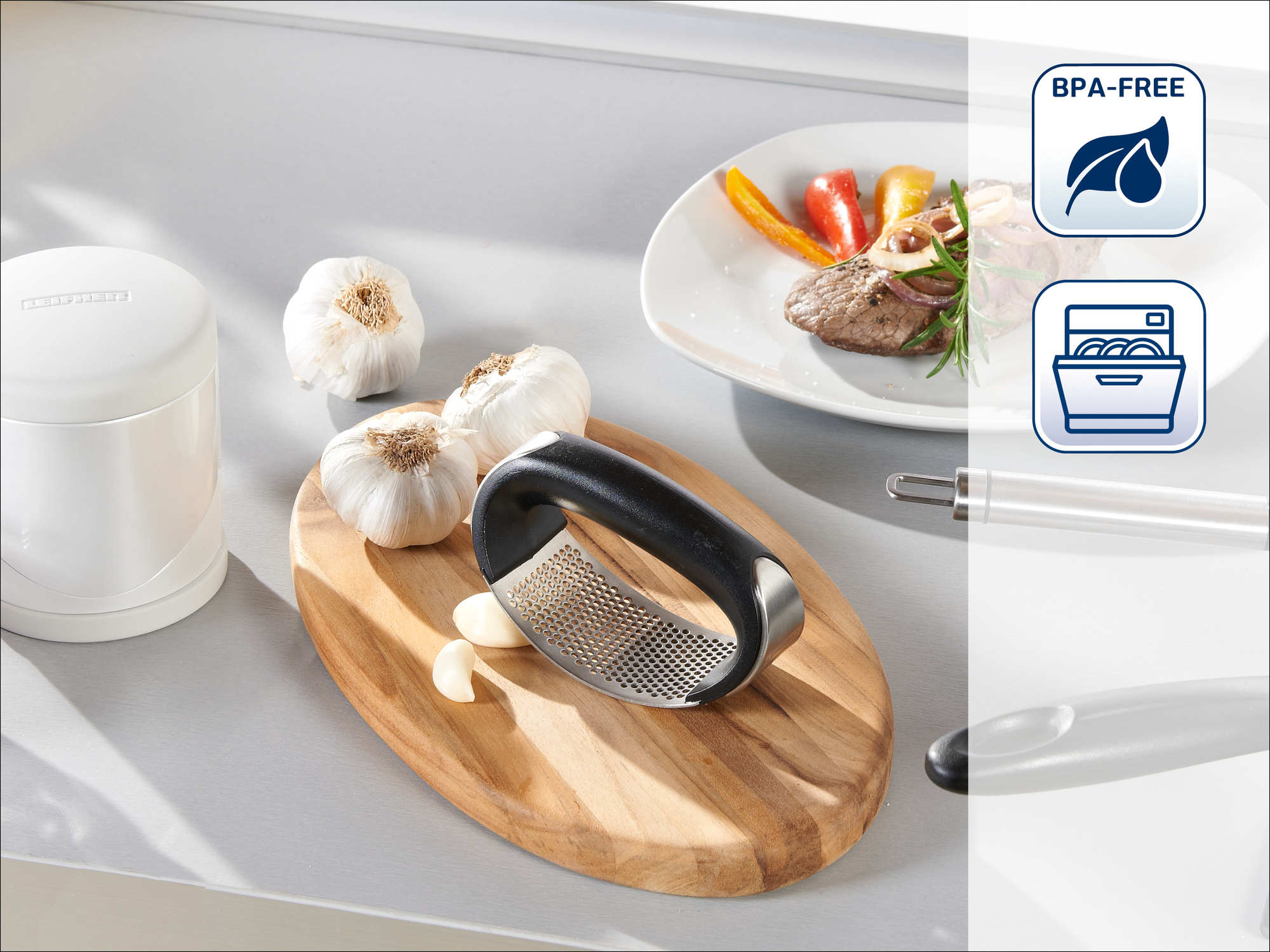 Praska do czosnku na drewnianej desce ze ząbkami czosnku, główkami i talerzem jedzenia. Biały pojemnik po lewej.