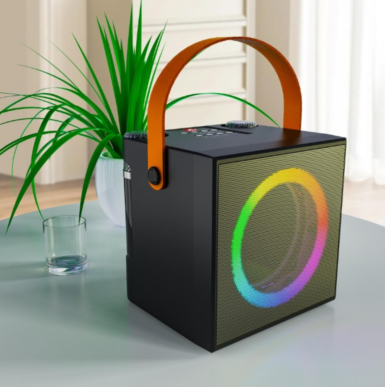 Schwarzer Lautsprecher mit orangefarbenem Griff, eine grüne Pflanze und ein Glas auf einem Tisch. Lautsprecher hat ein Regenbogenlicht.