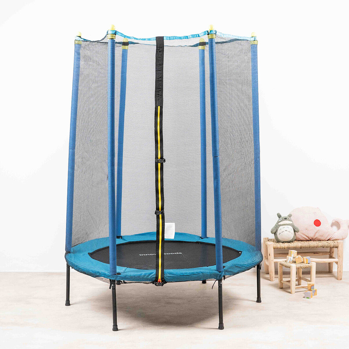 Un trampolín azul con red de seguridad está sobre un suelo claro. Un pequeño banco con juguetes y un peluche está cerca.