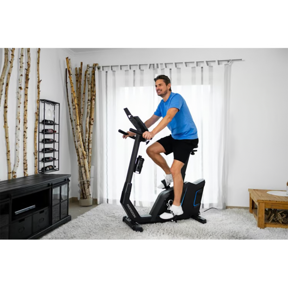 Un homme fait de l'exercice sur un vélo d'appartement dans une salle de sport à domicile. Il porte une chemise bleue et un short noir. Un meuble noir et des branches décoratives sont visibles.