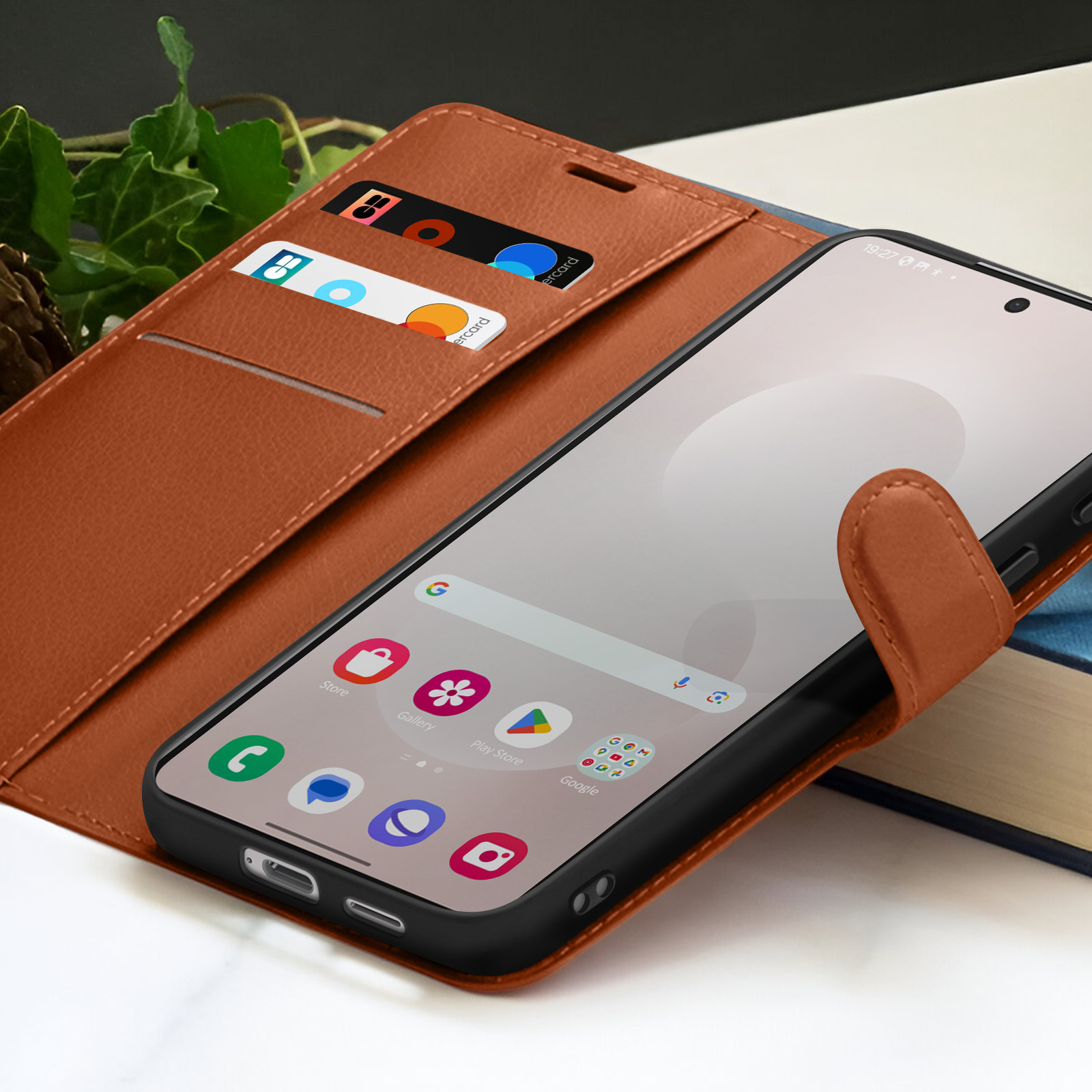 Een bruine leren telefoonhoes met een smartphone erin, met apps. Creditcards in de hoes. Een boek op de achtergrond.