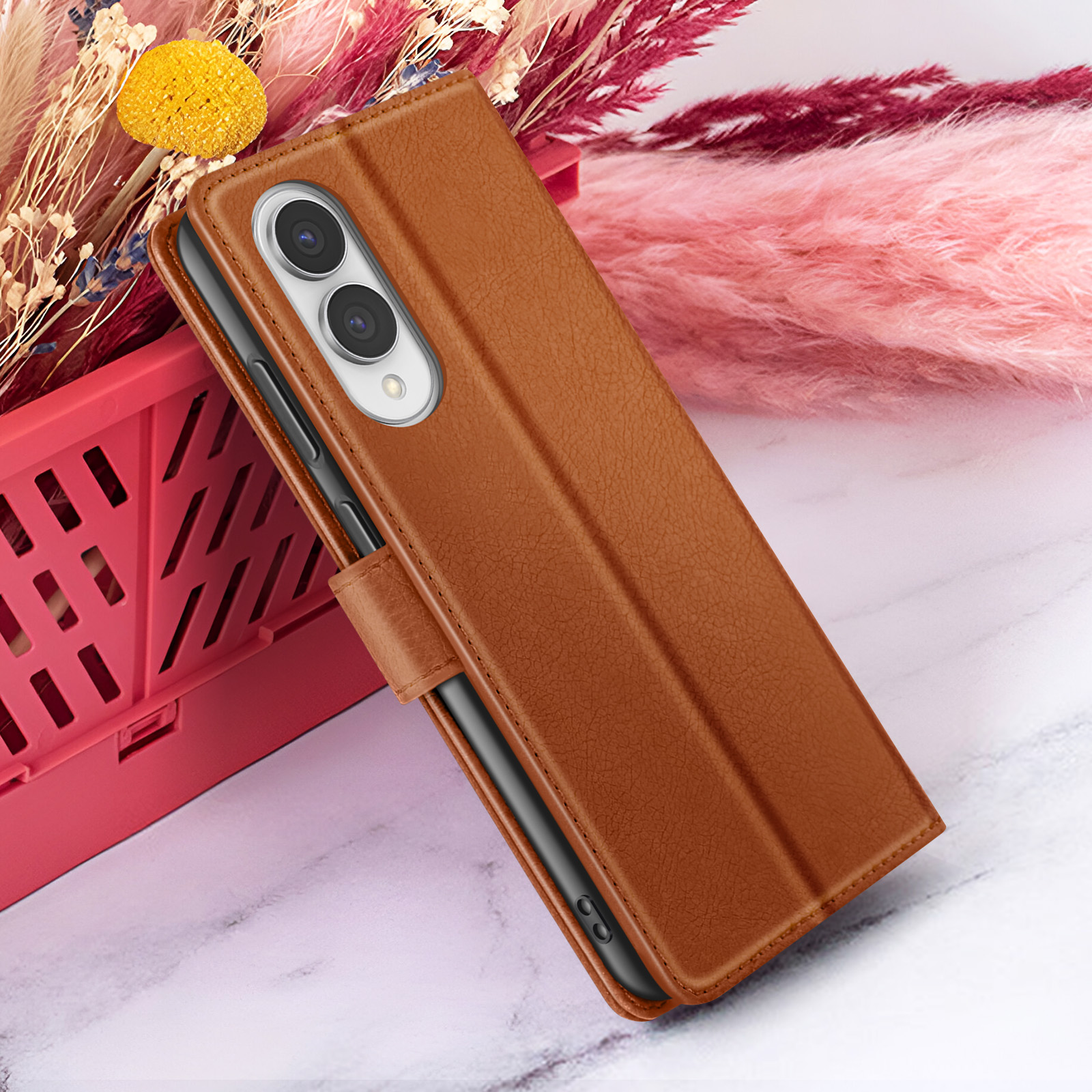 Bruine leren telefoonhoes met telefoon erin, tegen roze bloemen en rode mand op marmeren oppervlak.