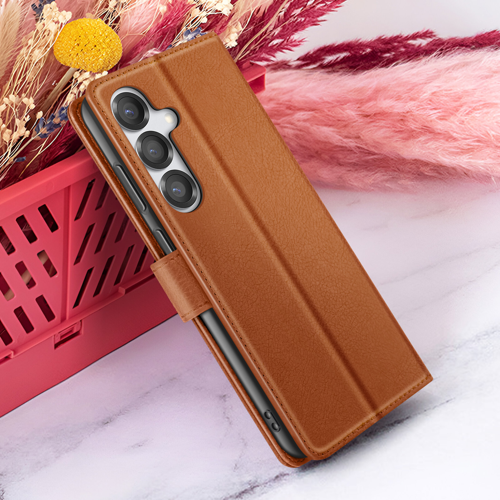 Een bruine leren telefoonhoes met een telefoon erin ligt op een marmeren oppervlak. Achtergrond heeft roze en rode bloemen.