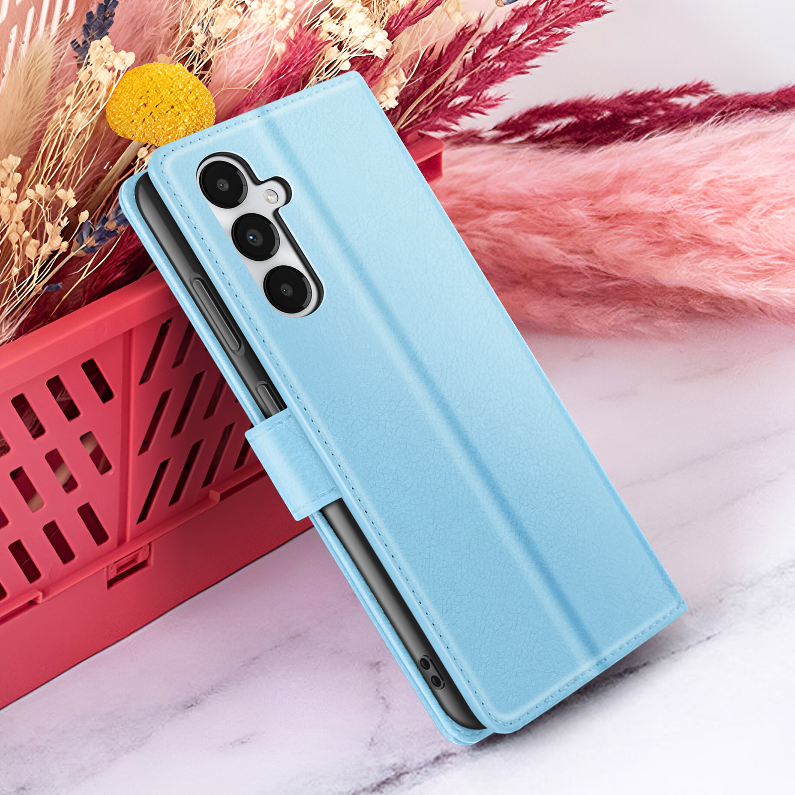 Un étui de téléphone bleu clair avec trois caméras est posé sur un panier rouge. Derrière, des fleurs séchées roses et beiges.