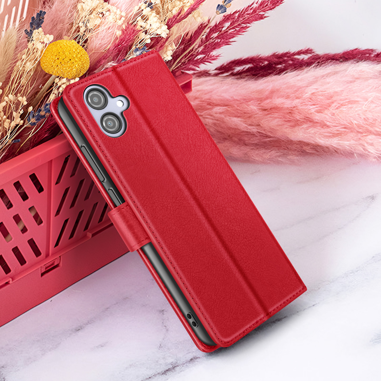 Étui de téléphone rouge avec caméra, soutenu par un panier rouge, avec des fleurs roses et beiges en arrière-plan.