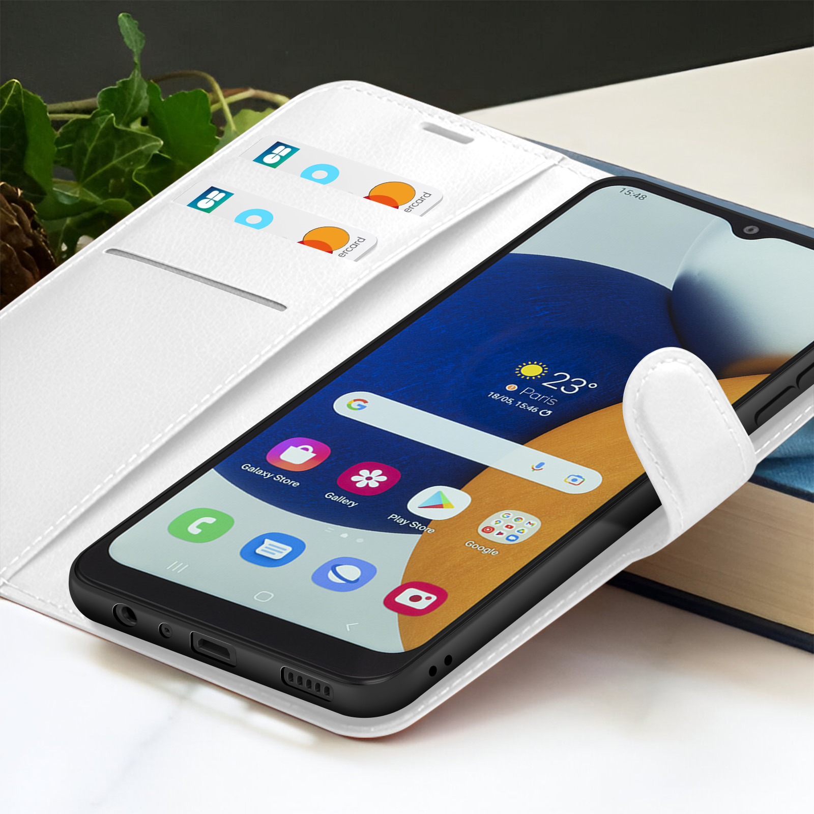 Un smartphone dans un étui blanc affiche l'écran d'accueil. Cartes de crédit visibles. Feuillage vert en arrière-plan.