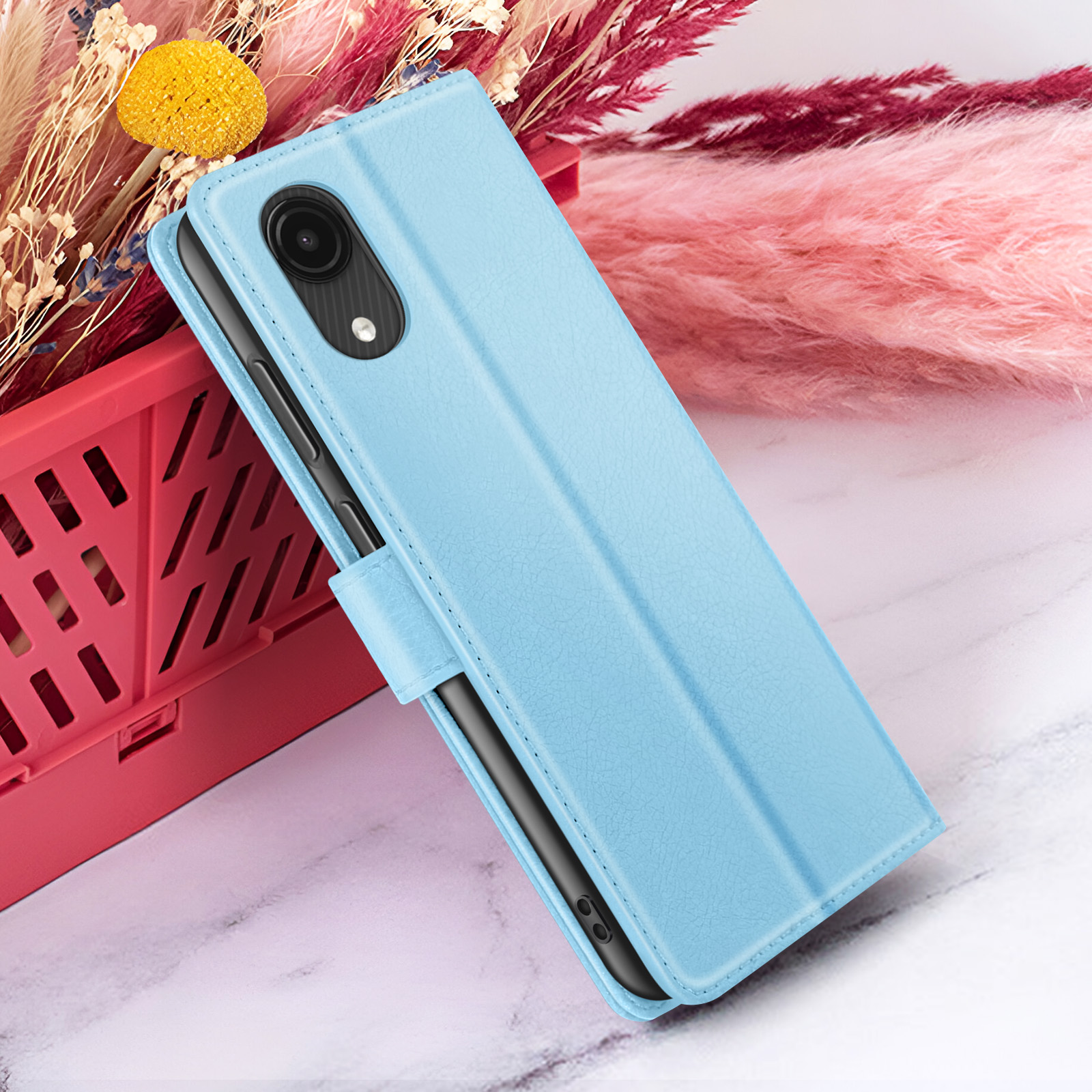 Una custodia per telefono azzurra è aperta, mostrando un telefono nero. È accanto a fiori rosa e un cestino rosso.