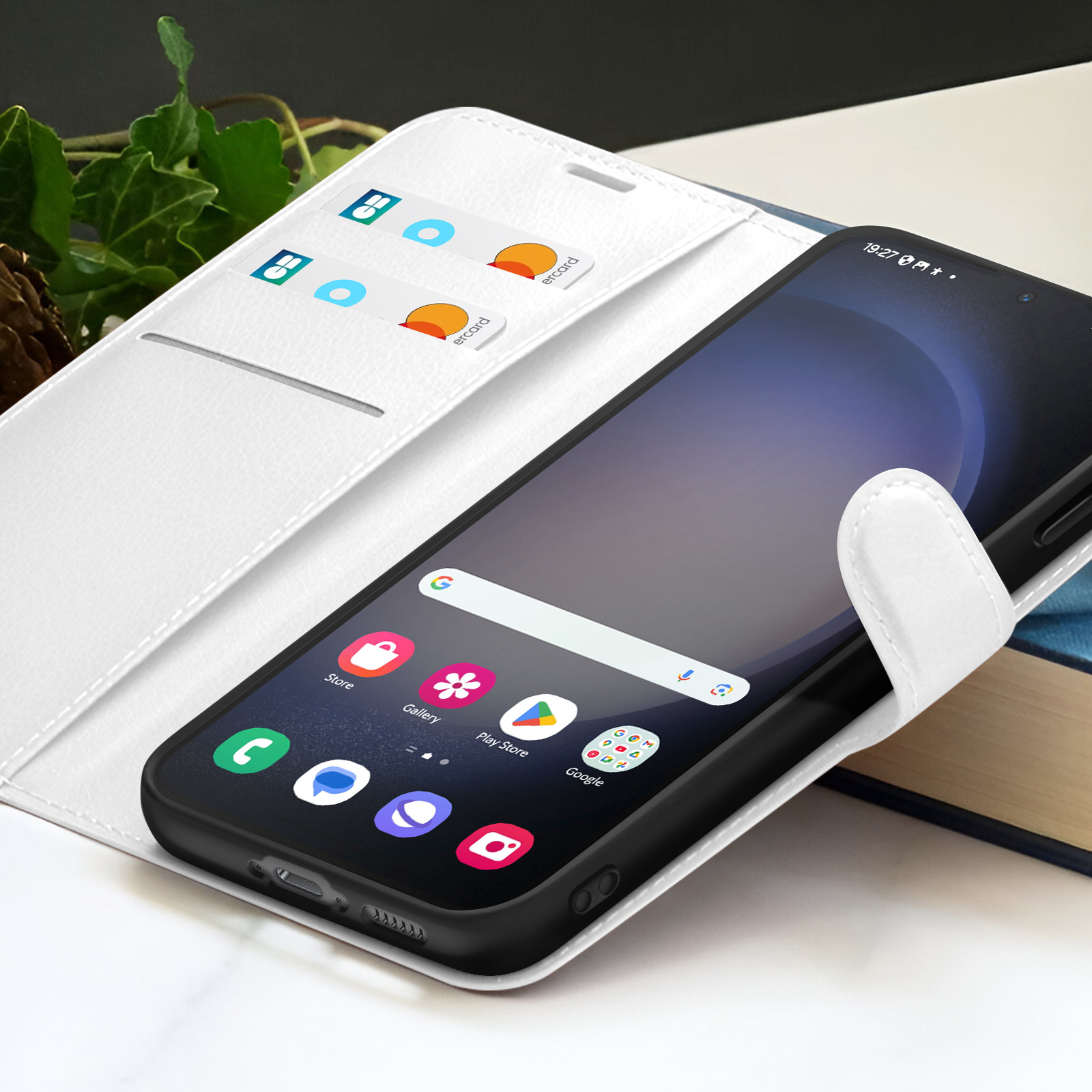 Zwarte smartphone in witte hoes, pictogrammen op scherm. Twee kaarten zichtbaar. Klimop op de achtergrond. Boek rechts.