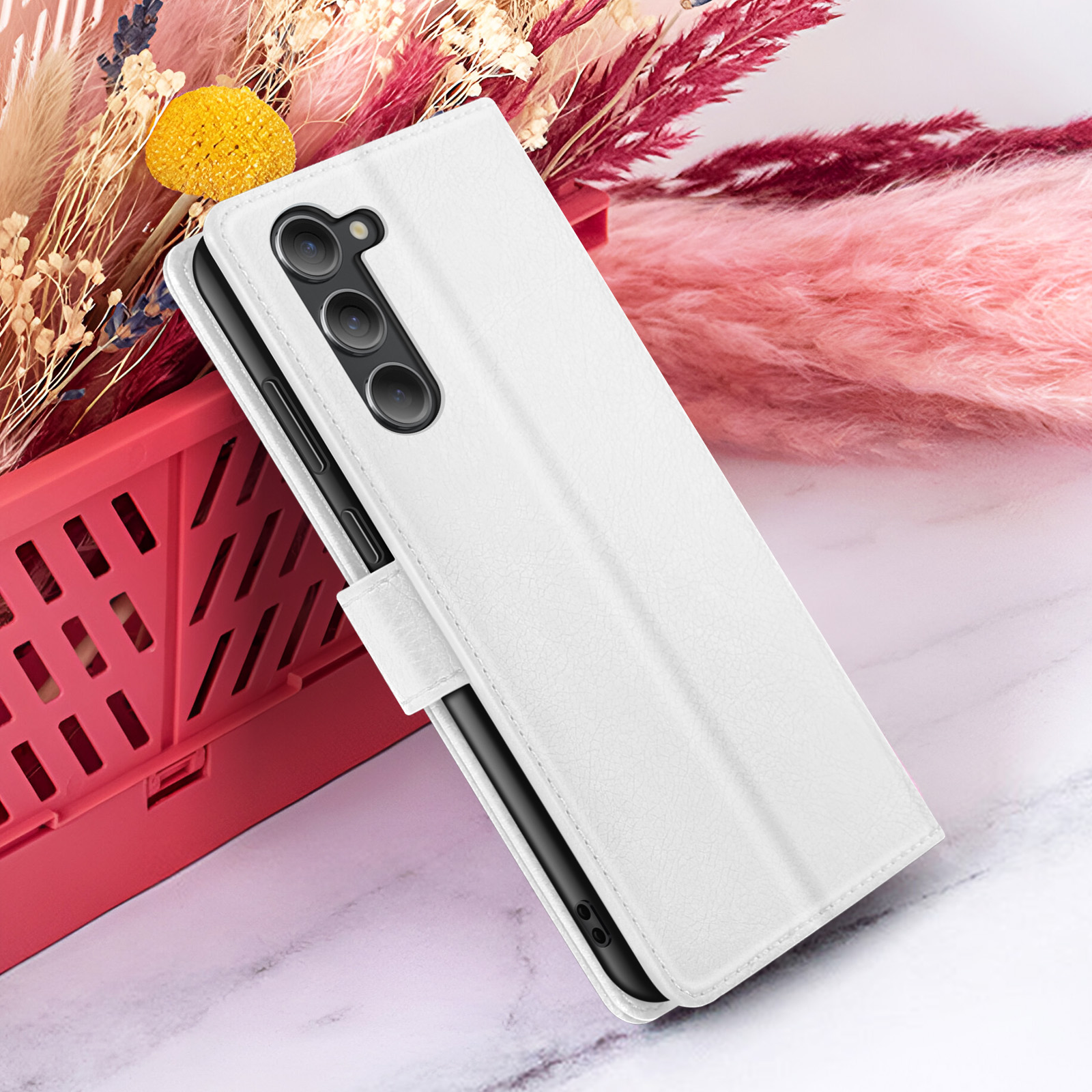 Een witte telefoonhoes ligt op een marmeren oppervlak met gedroogde bloemen en een rode mand op de achtergrond.