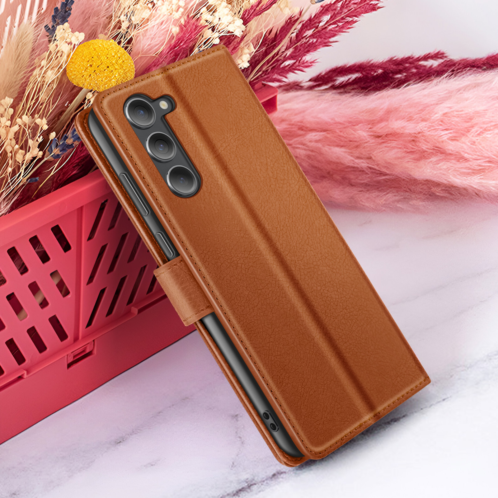 Bruine leren telefoonhoes met een telefoon erin, naast roze en witte bloemen in een rode mand.