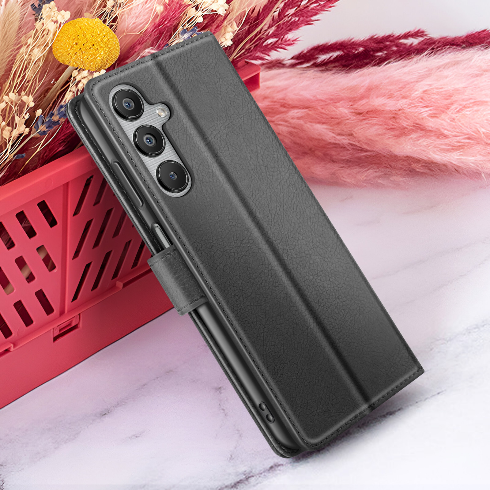 Un étui de téléphone noir avec trois lentilles d'appareil photo est ouvert. Derrière, des fleurs roses dans un panier rouge, sur du marbre.