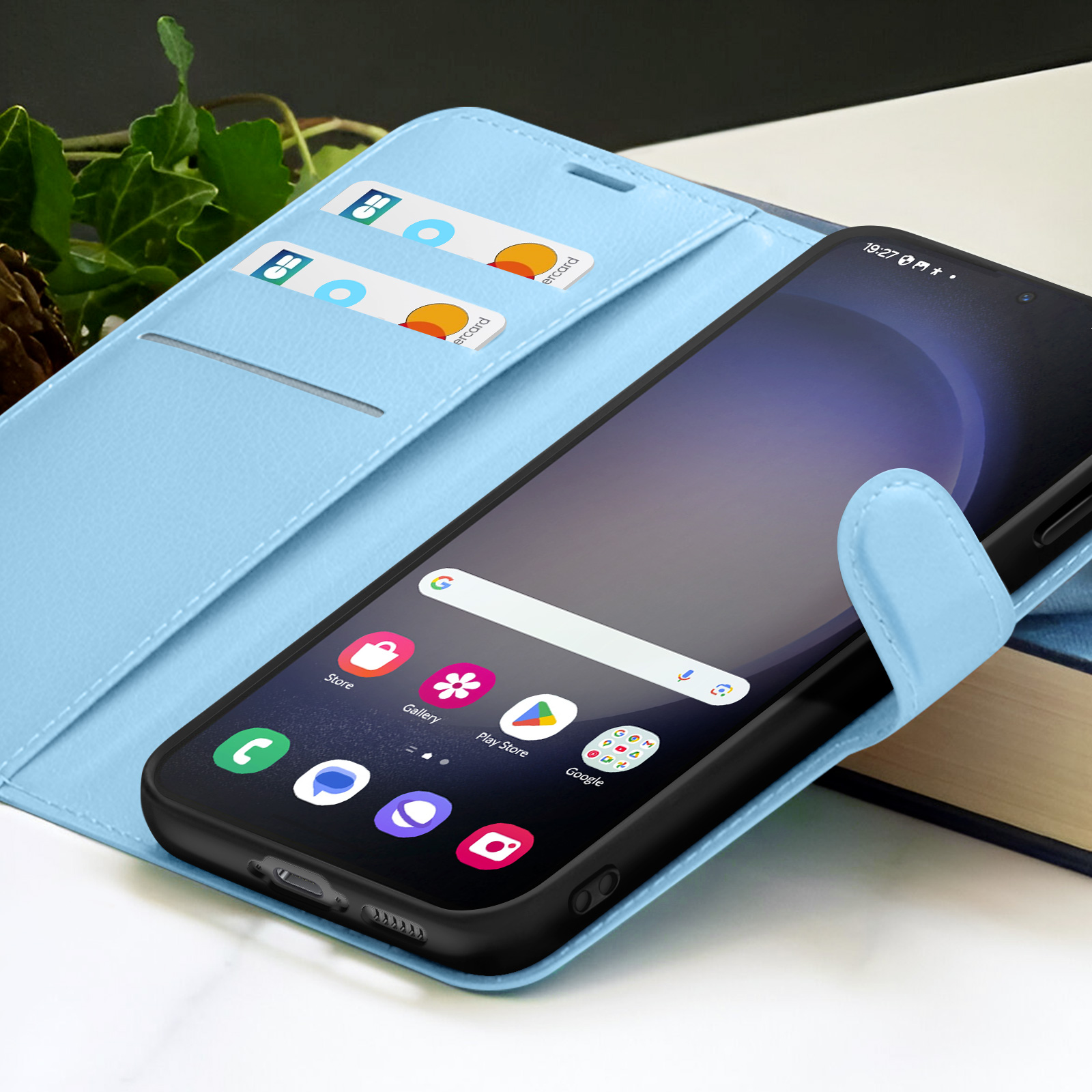 Un smartphone dans un étui bleu clair, avec des fentes pour cartes de crédit. L'écran affiche des icônes d'applications. Arrière-plan avec feuillage et livre.