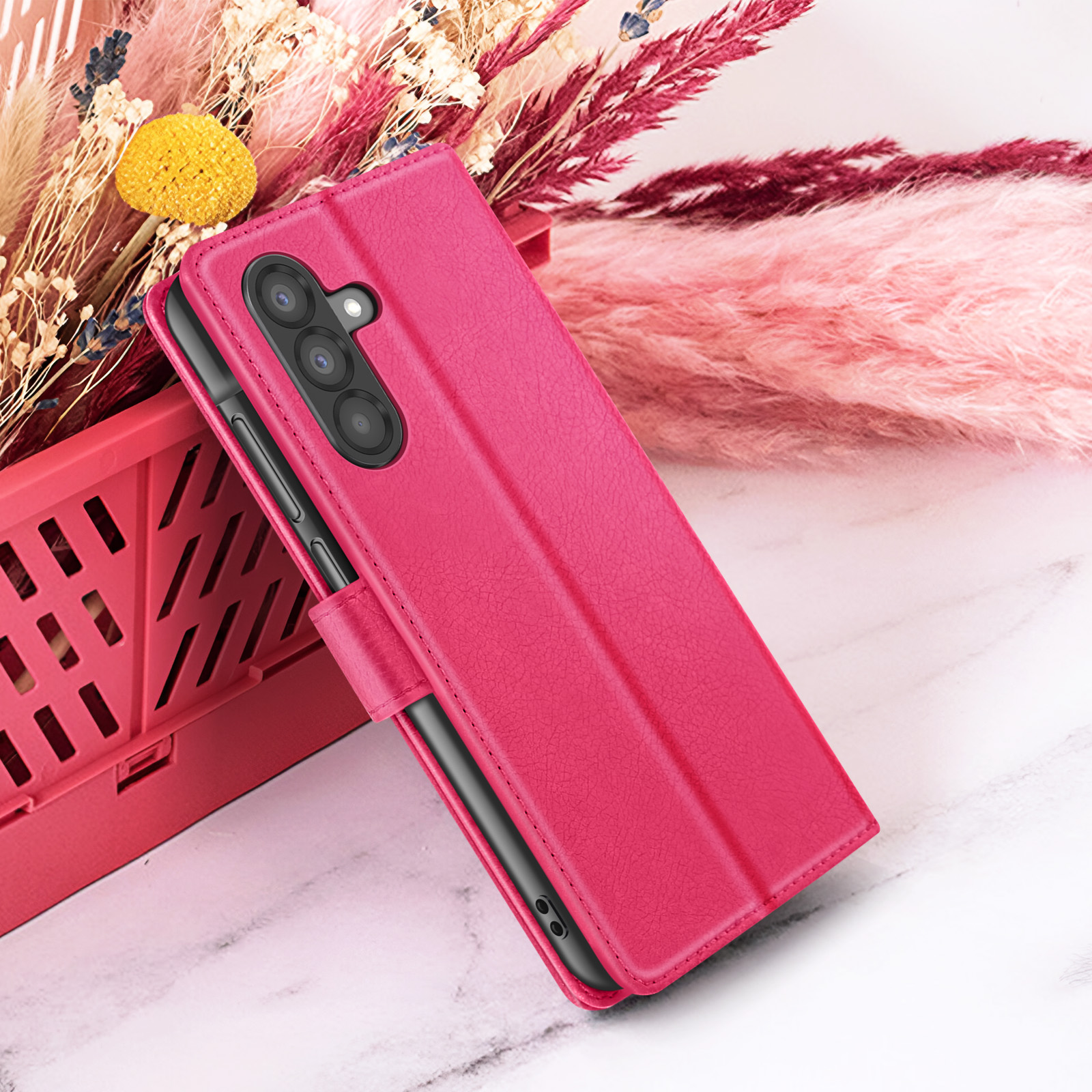 Een fuchsia telefoonhoesje op een marmeren oppervlak, met gedroogde bloemen op de achtergrond. De hoes heeft een uitsparing voor de camera.
