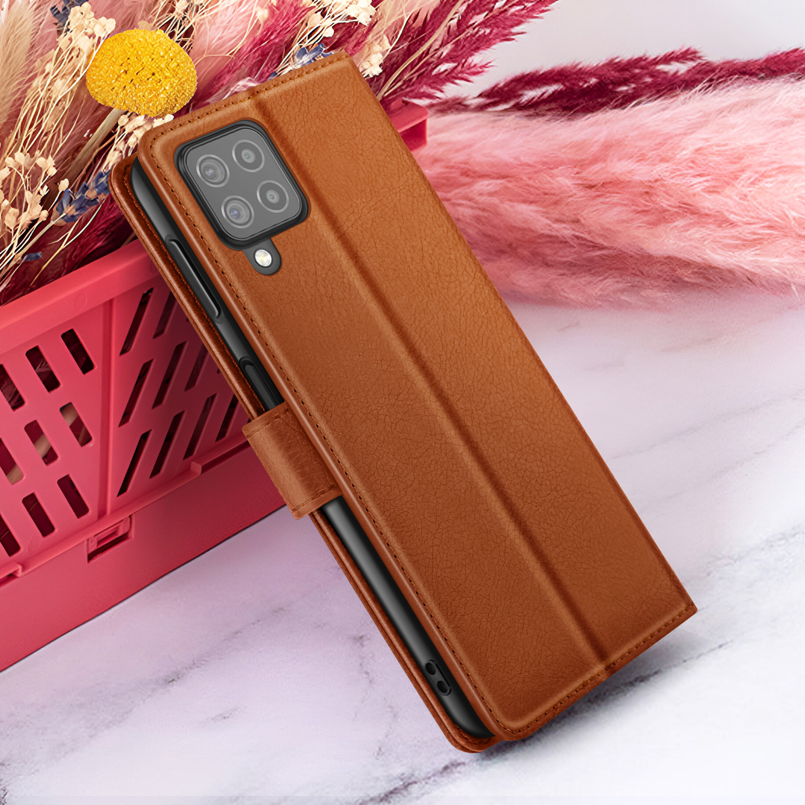 Bruine leren telefoonhoes met camera en flap. Op de achtergrond roze bloemen en een rode container.