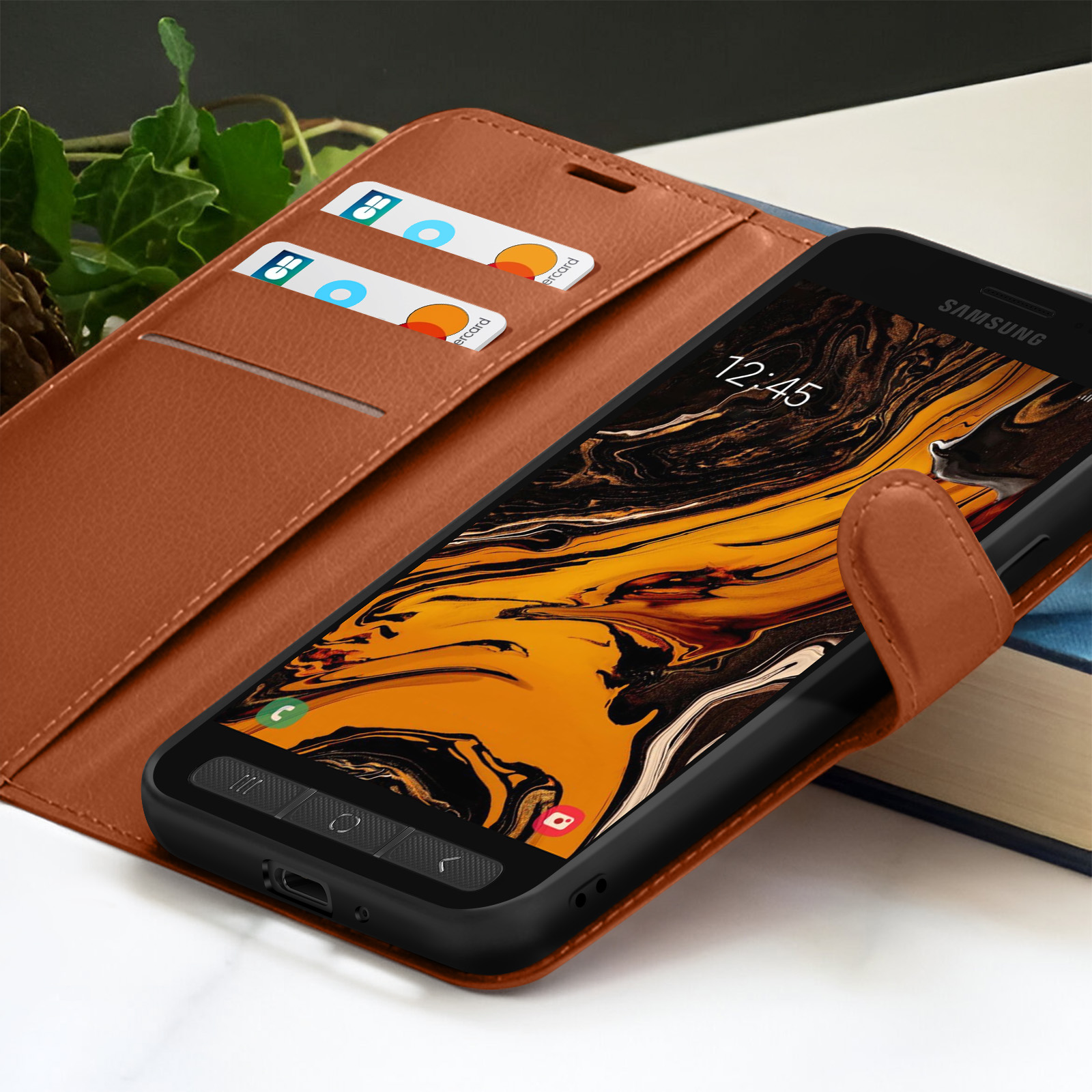 Téléphone Samsung dans un étui en cuir marron, avec des cartes dans les fentes. L'écran affiche 12:45. Fond : livre et feuilles.