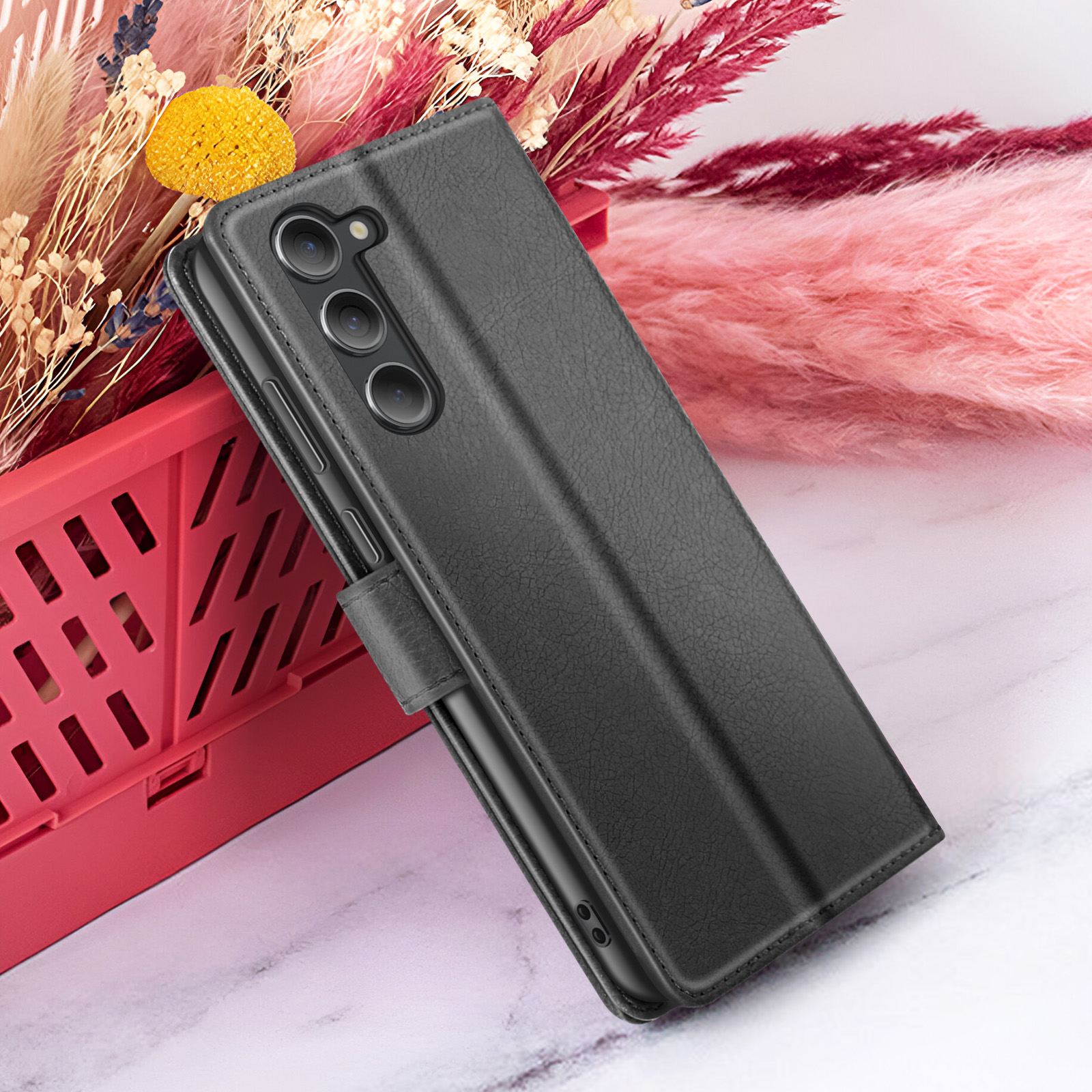 Une coque de téléphone noire avec une surface texturée, la caméra du téléphone est visible. Elle est placée près d'une décoration rose et rouge.