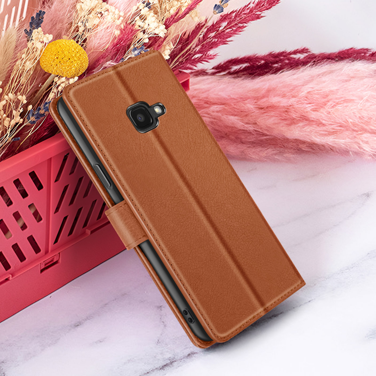 Un étui de téléphone en cuir marron est ouvert, affichant un téléphone. Fleurs séchées et décor rose duveteux en arrière-plan.