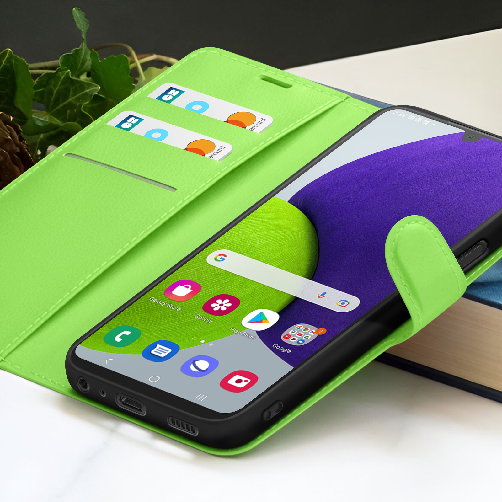 Een smartphone in een limoengroene hoes toont apps. Twee creditcards zitten in de hoes. De achtergrond bevat een boek en groene bladeren.