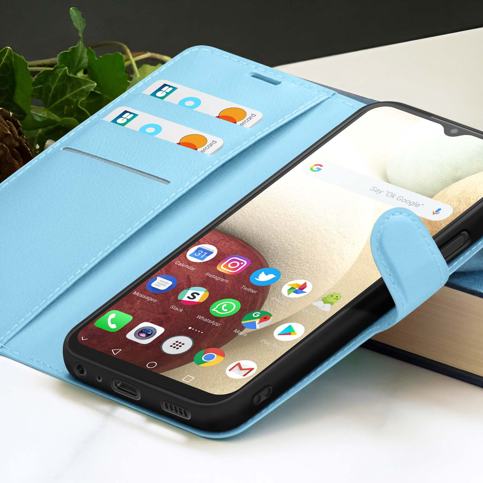 Smartphone in een lichtblauwe hoes met kaarten. De telefoon toont app-iconen. Achtergrond is klimop en boeken.
