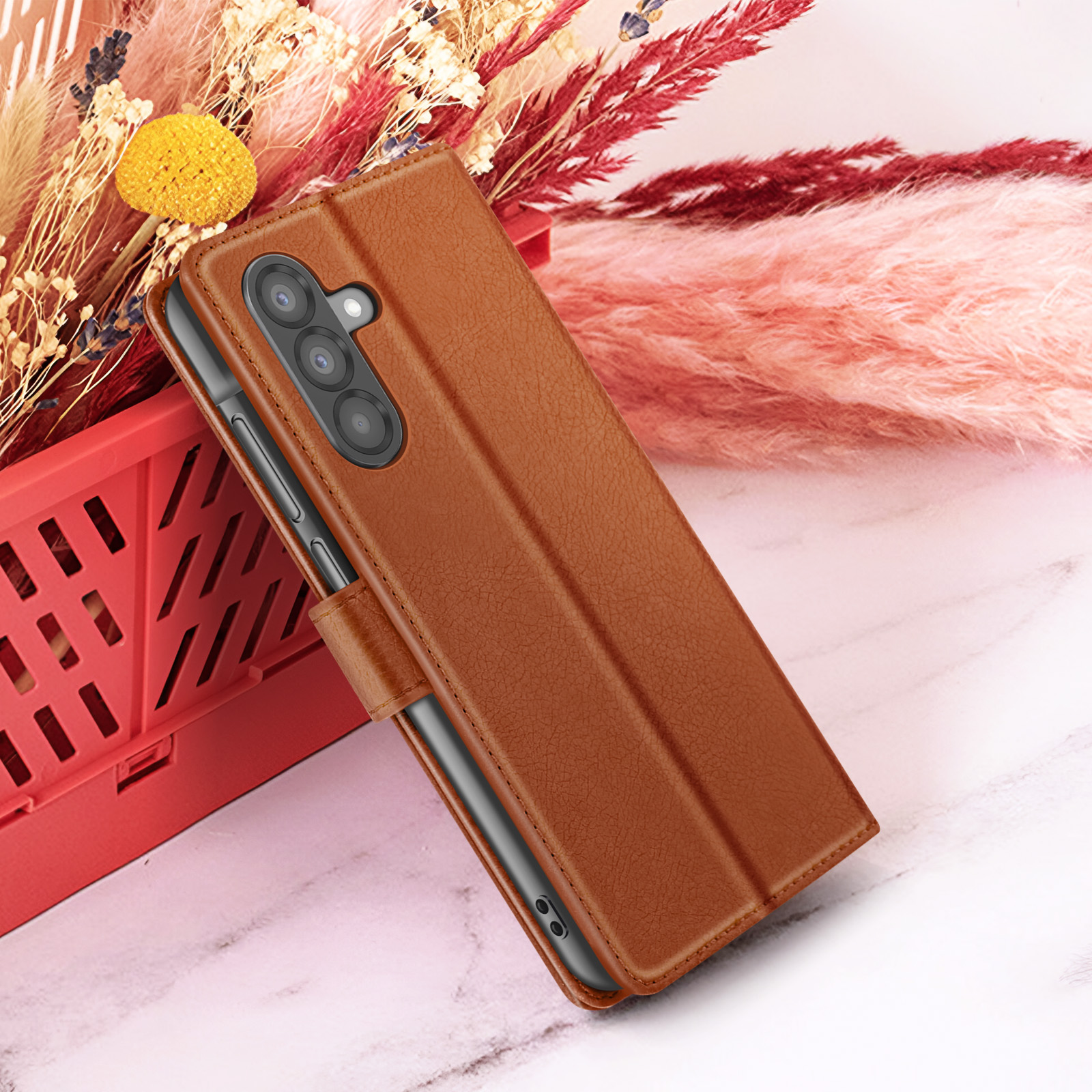 Un étui de téléphone en cuir marron avec un téléphone à l'intérieur, placé à côté de fleurs dans un conteneur rouge, sur une surface en marbre.