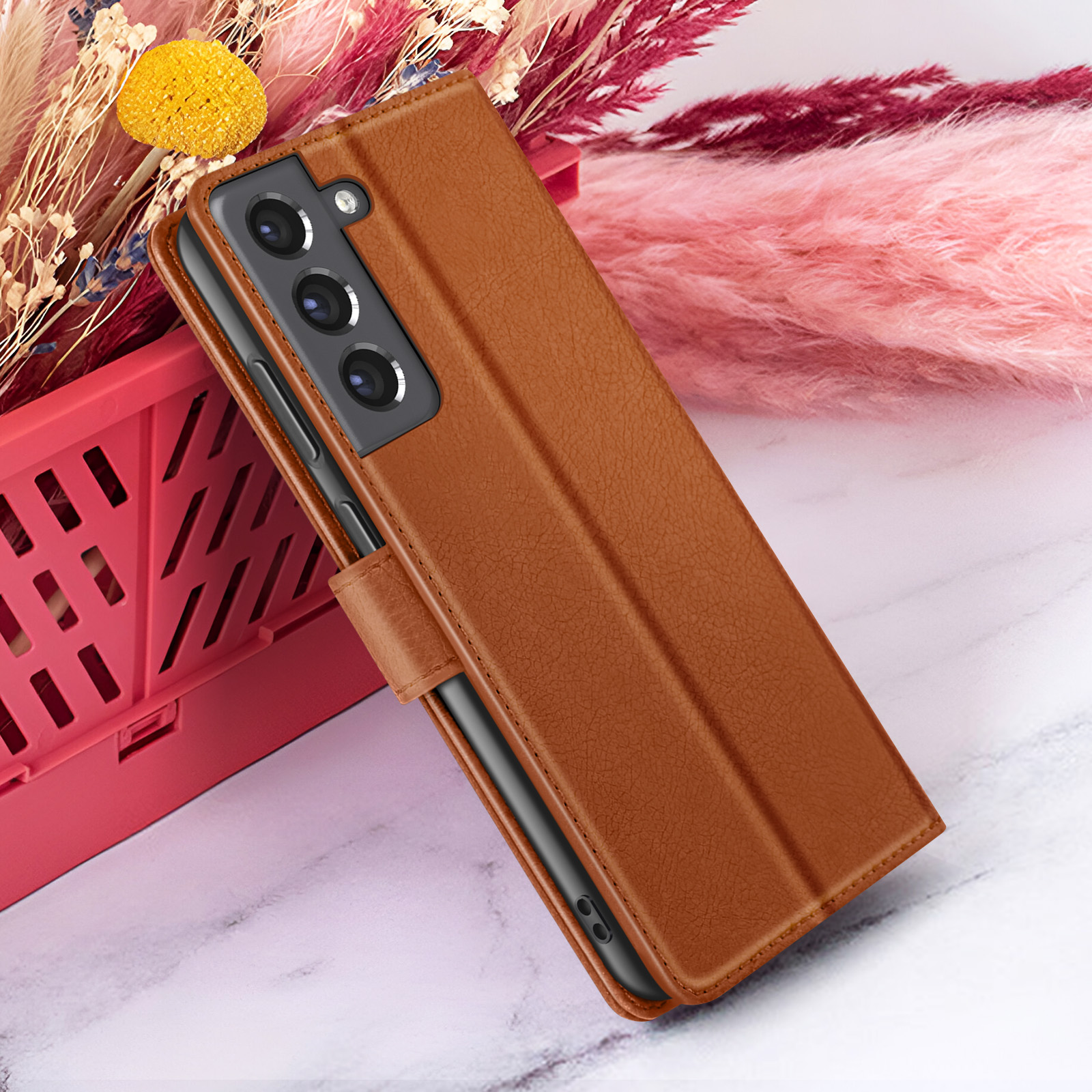 Bruine leren telefoonhoes met telefoon erin, rustend tegen bloemen in een rode mand en roze achtergrond.