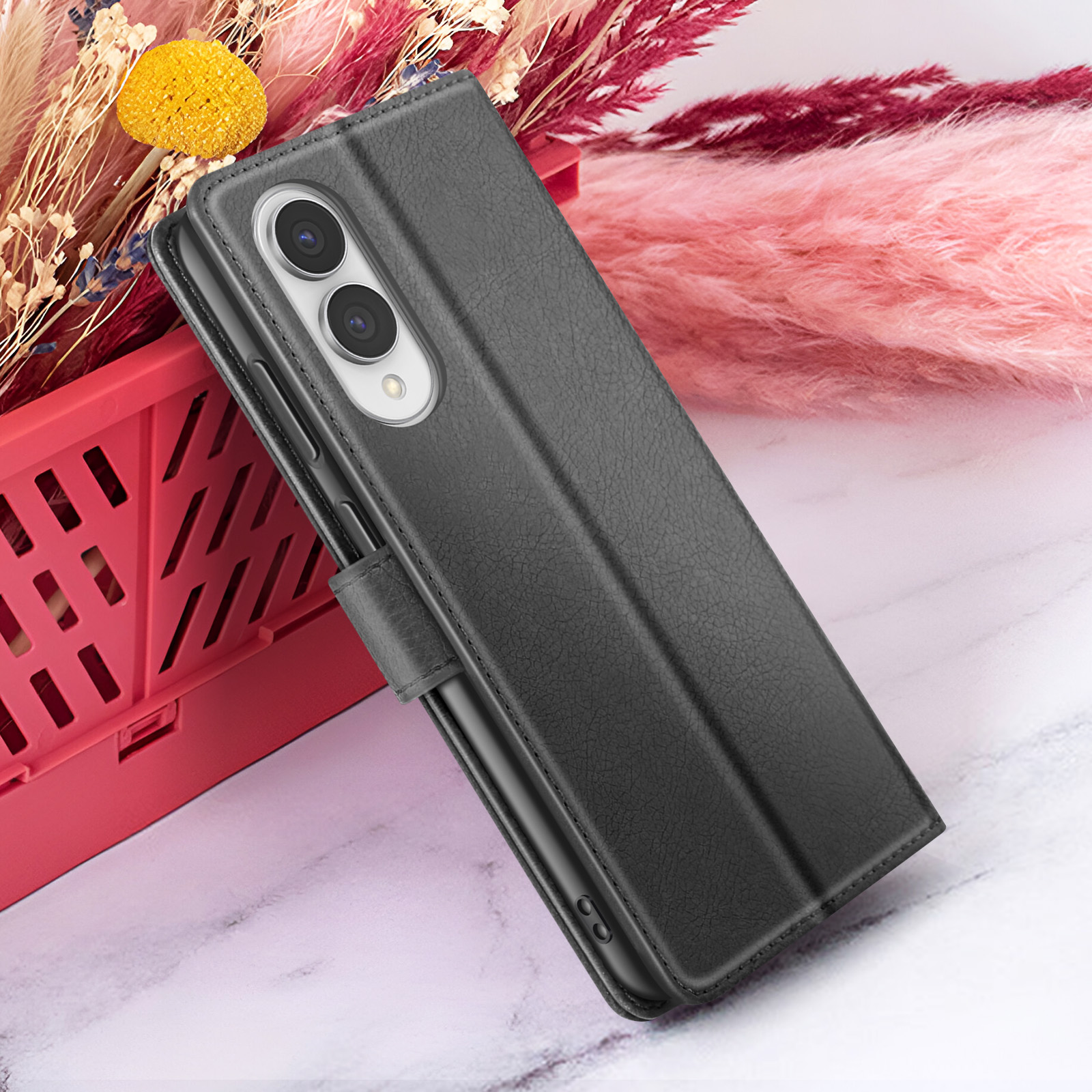 Un étui de téléphone noir repose contre un panier rouge avec des fleurs sur une surface en marbre. L'étui a deux caméras.
