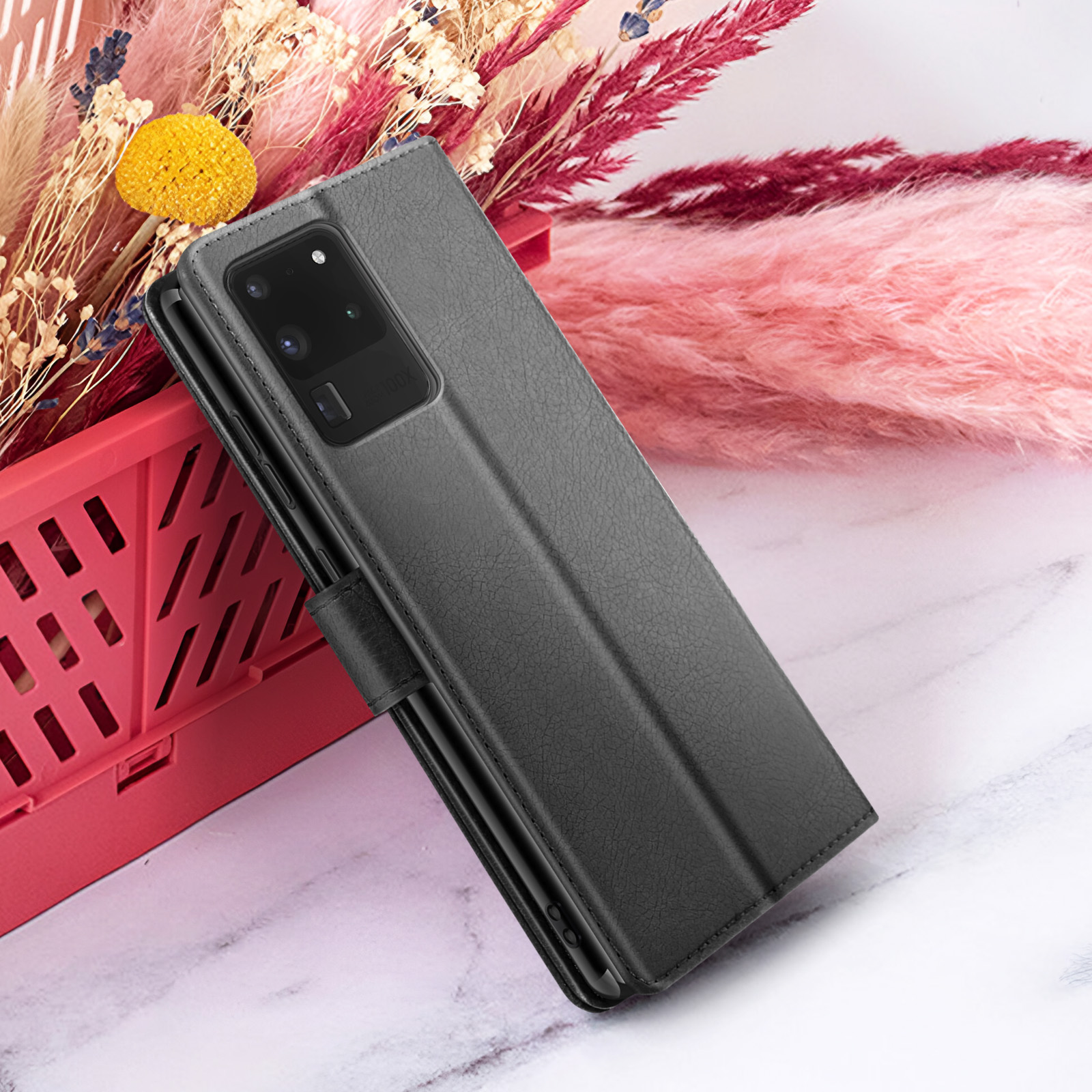Un étui de téléphone noir est présenté. Il est appuyé contre un panier rouge avec des fleurs et un fond rose et duveteux.