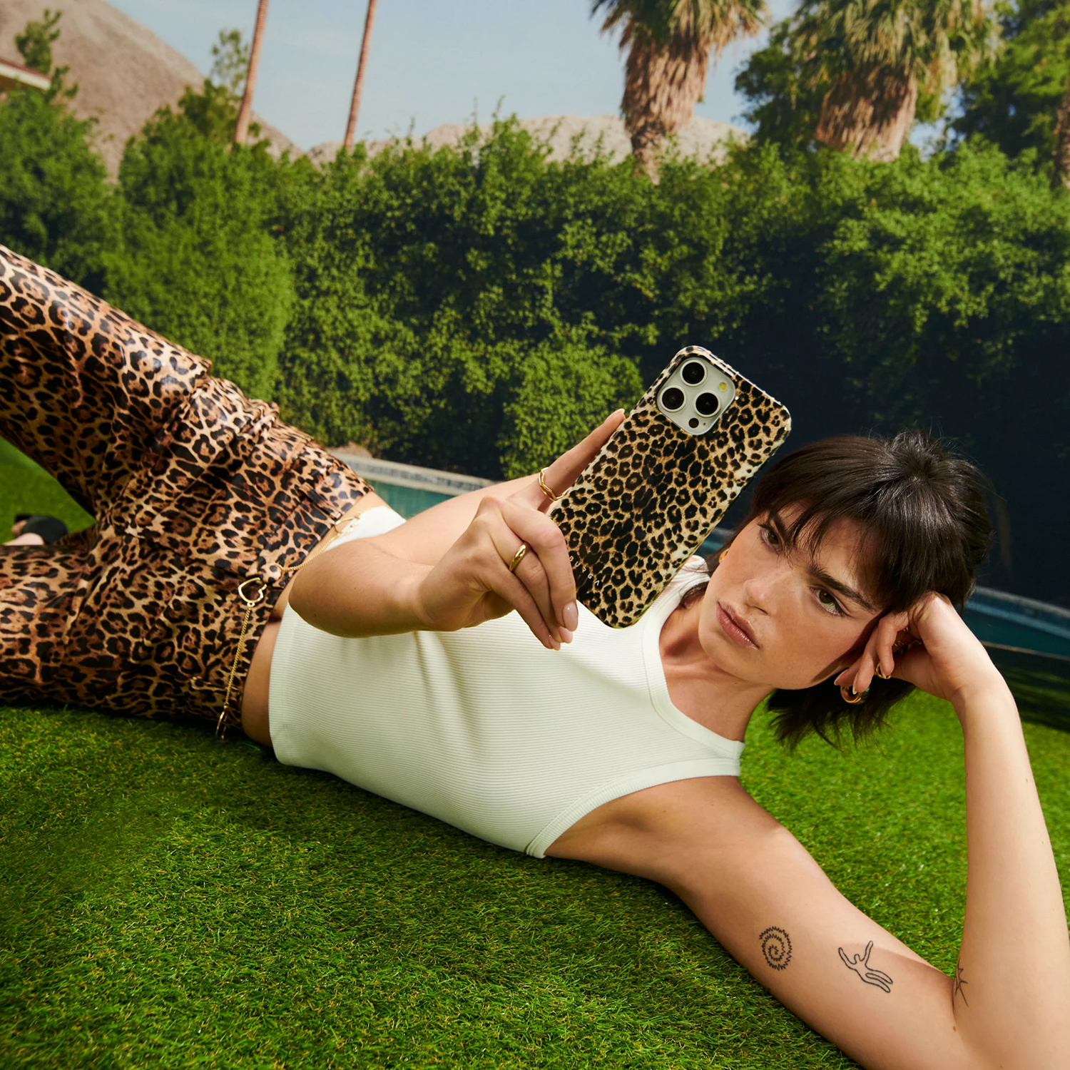 Une femme avec un étui de téléphone et un pantalon à imprimé léopard est allongée sur l'herbe, regardant le téléphone. Des palmiers et des buissons verts en arrière-plan.